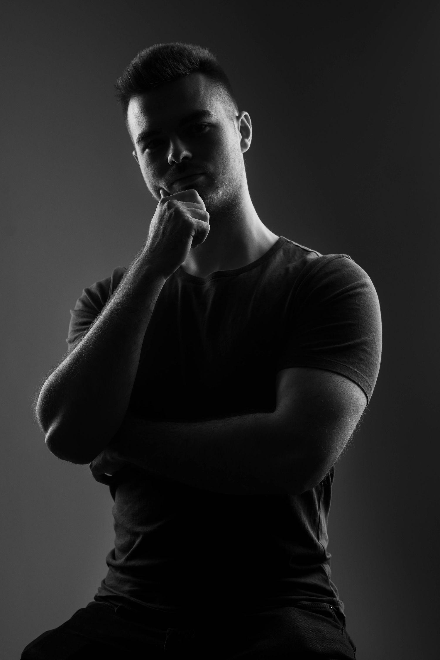 Dramatic black and white portrait of person in t-shirt with hand on chin against dark background.