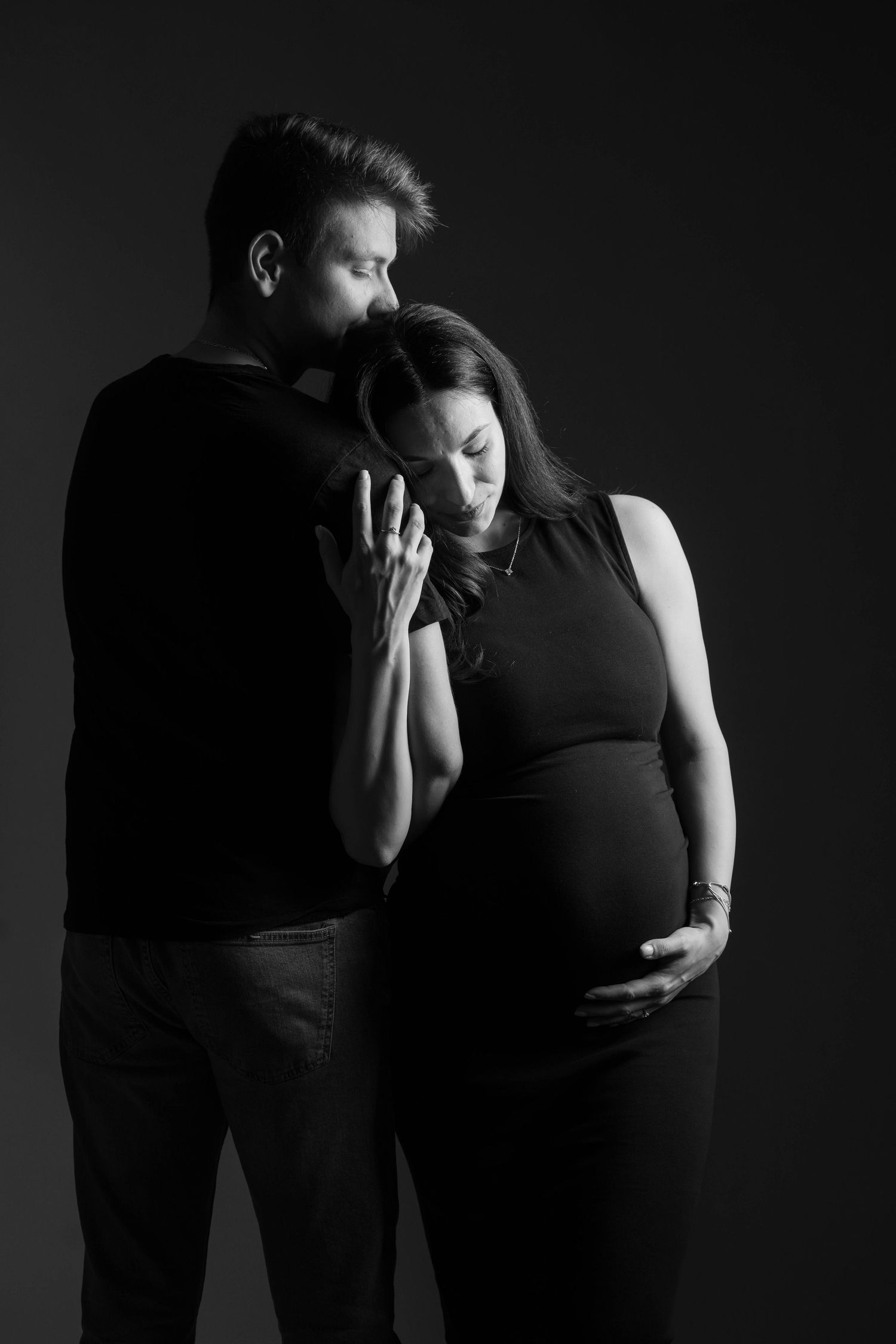 A dramatic black and white maternity photo shows a couple embracing in an intimate pose against a dark background.