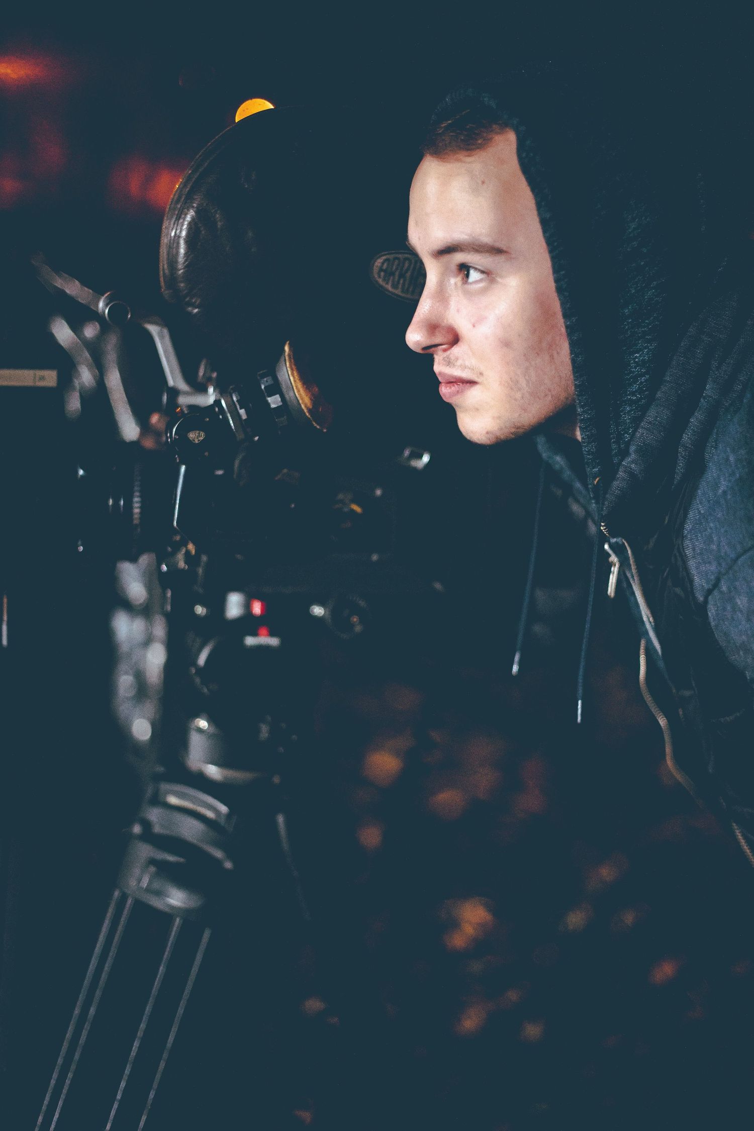 Camera operator in dark setting working with professional film equipment and tripod in profile view.