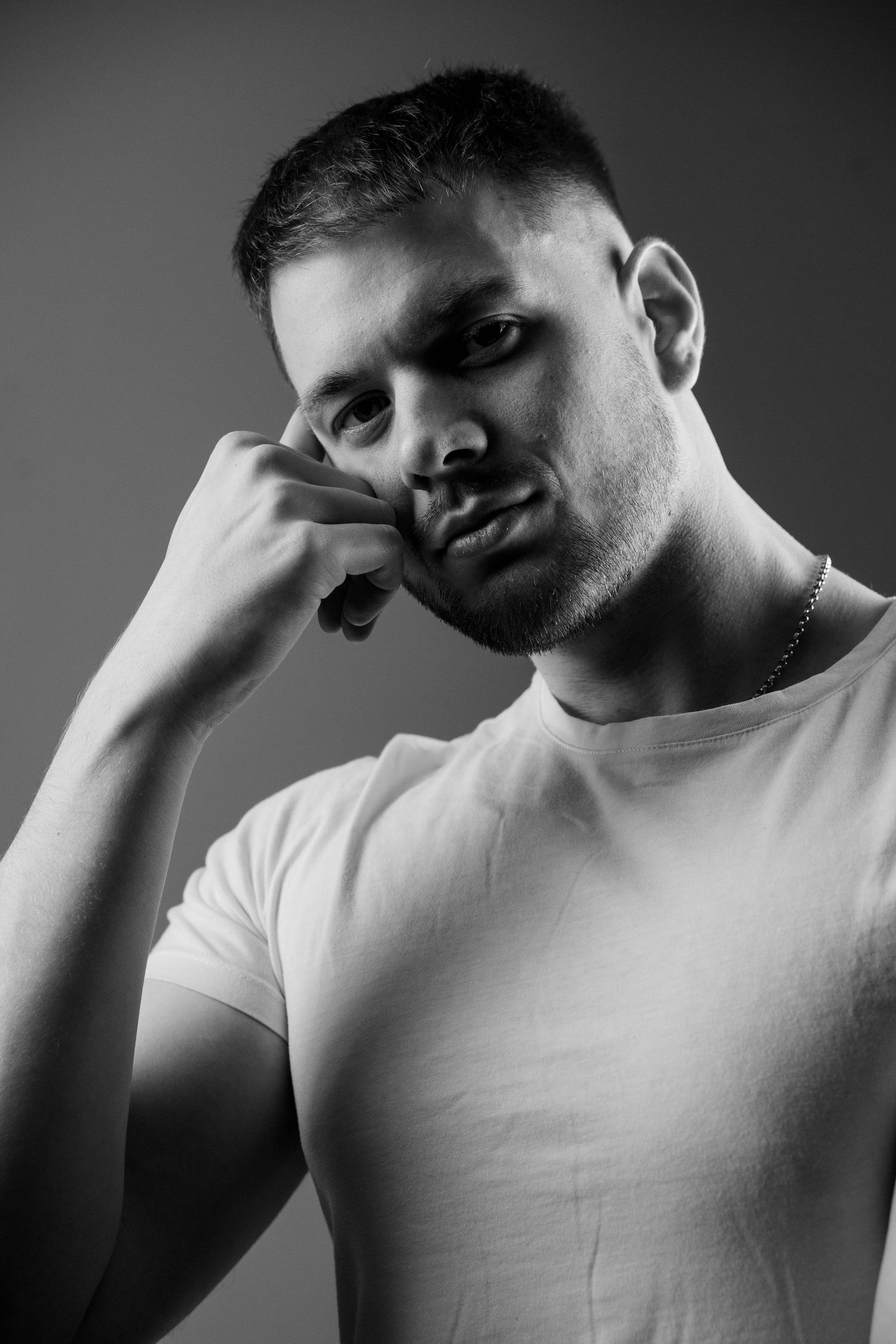 Black and white portrait of a person in white t-shirt with head resting thoughtfully on hand against dark background.