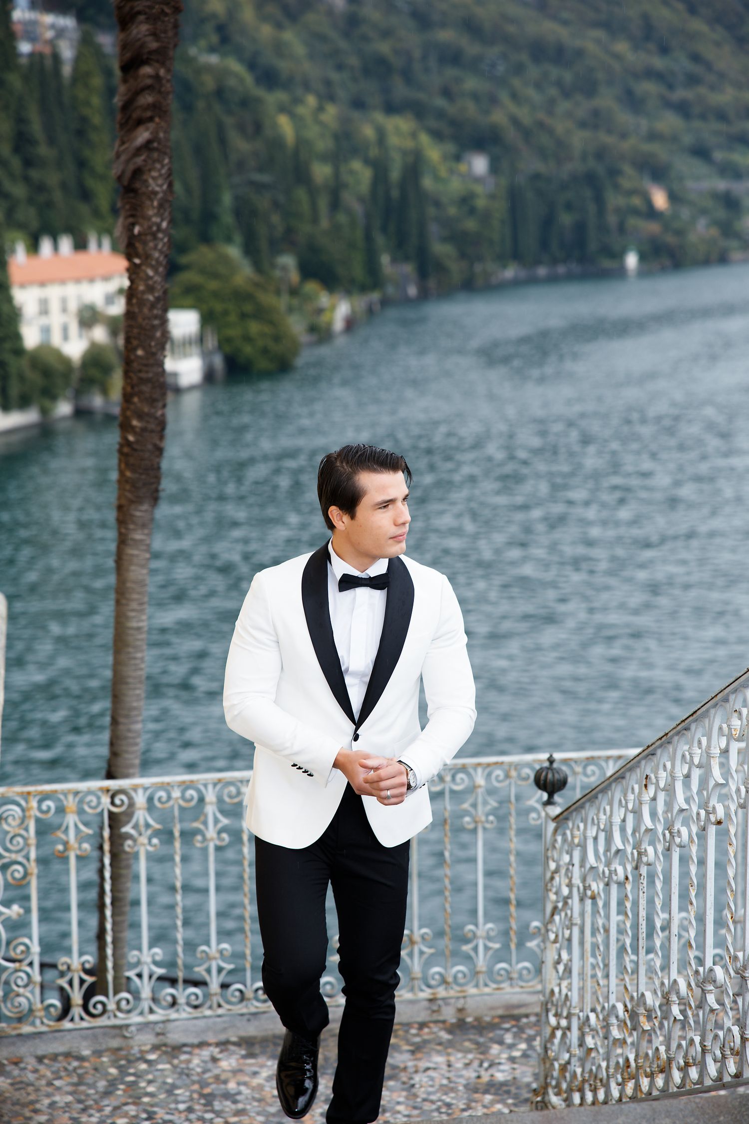 Groom in white tuxedo jacket poses on ornate balcony overlooking Italian lake scenery.