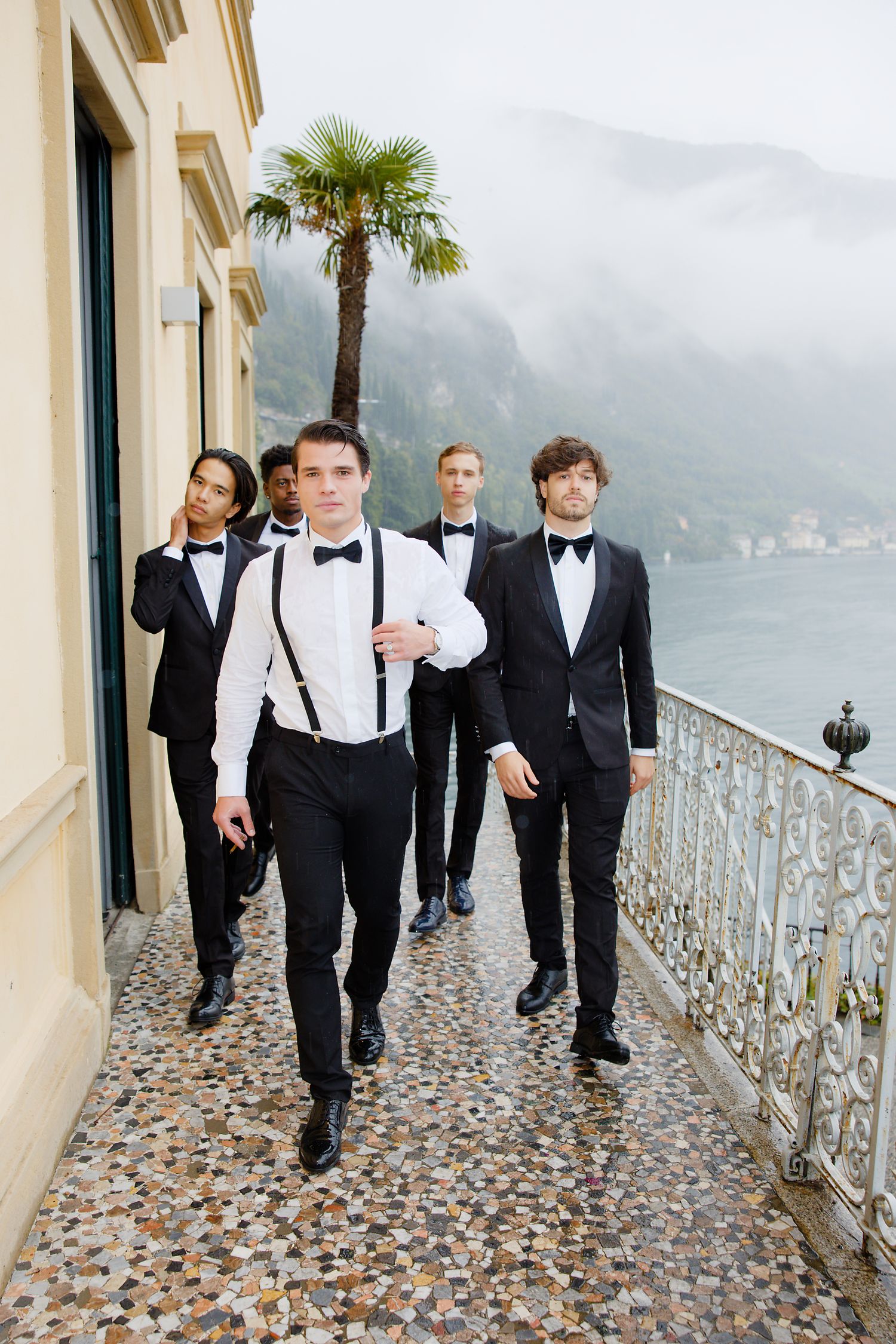 Groomsmen in formal attire walk along a scenic balcony overlooking a lake and mountains.