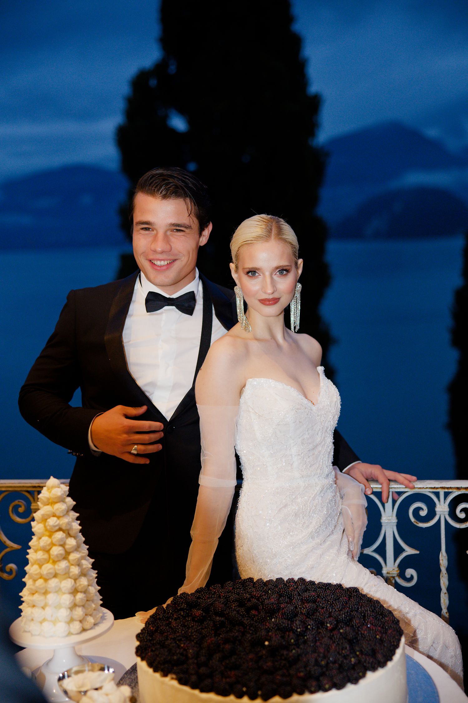 An elegant wedding reception setup with contrasting black and white cakes against a twilight Mediterranean backdrop.