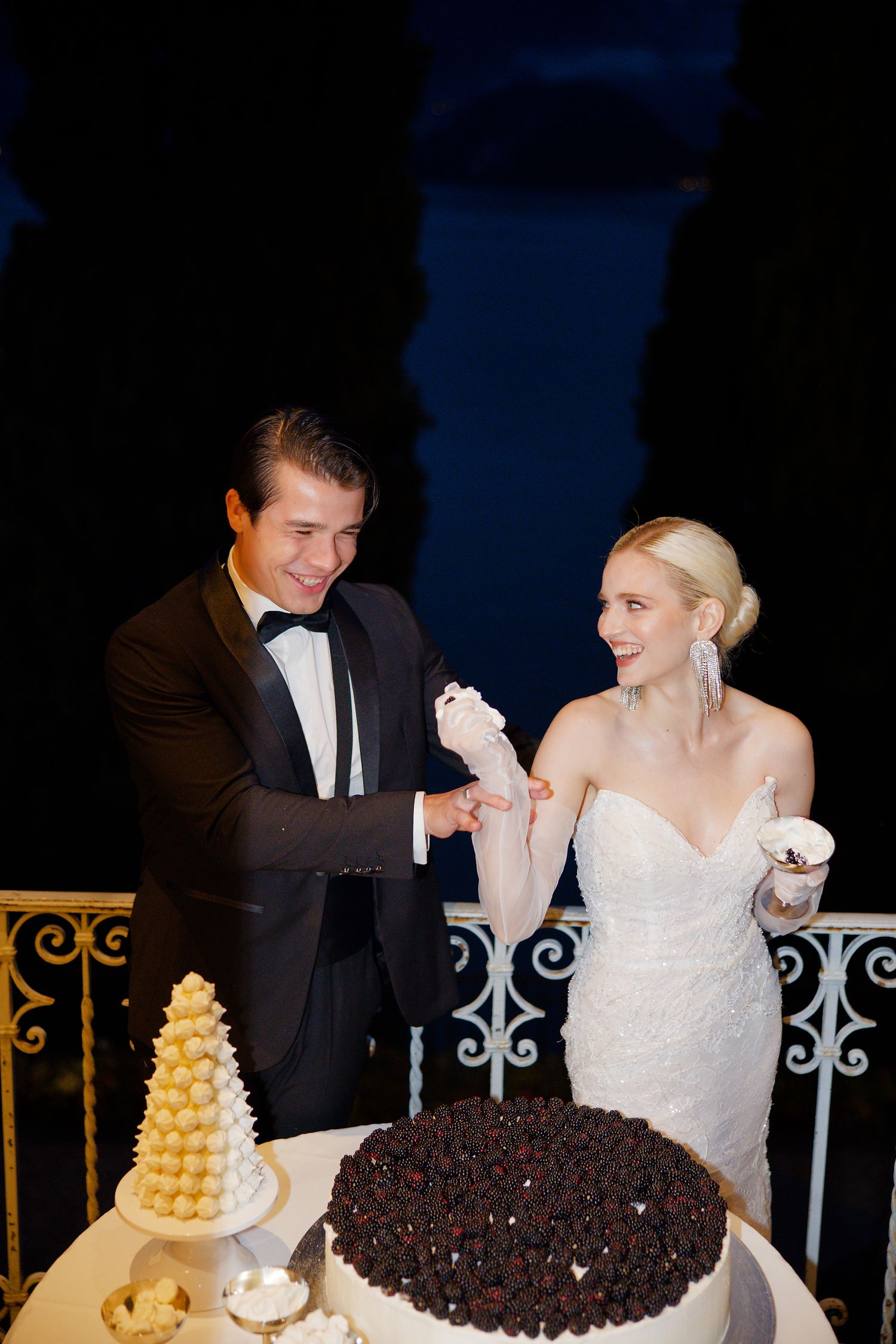 Elegant wedding couple cutting cake at nighttime reception on ornate balcony.