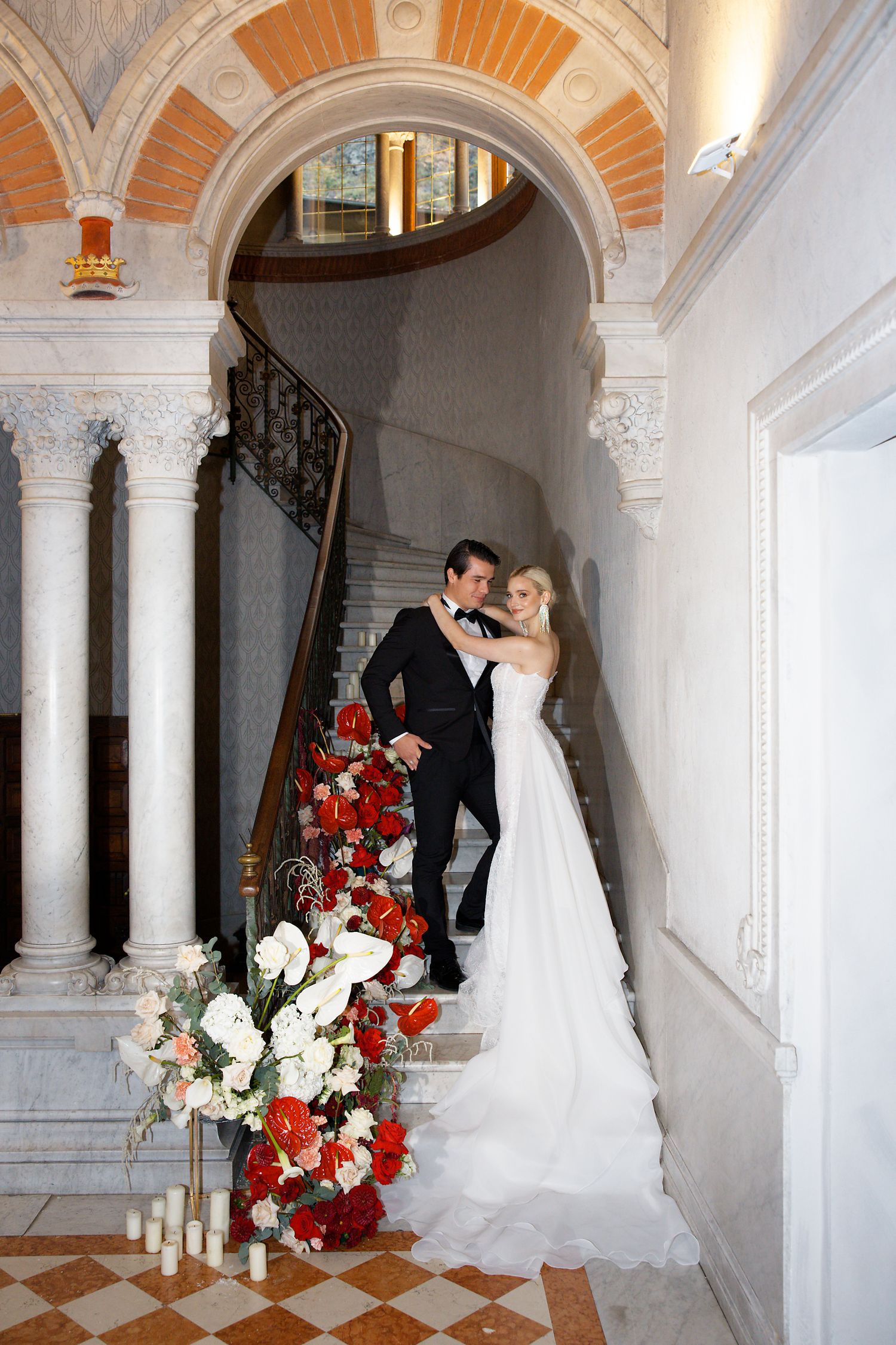 Wedding couple poses in elegant marble archway decorated with red and white floral arrangements.