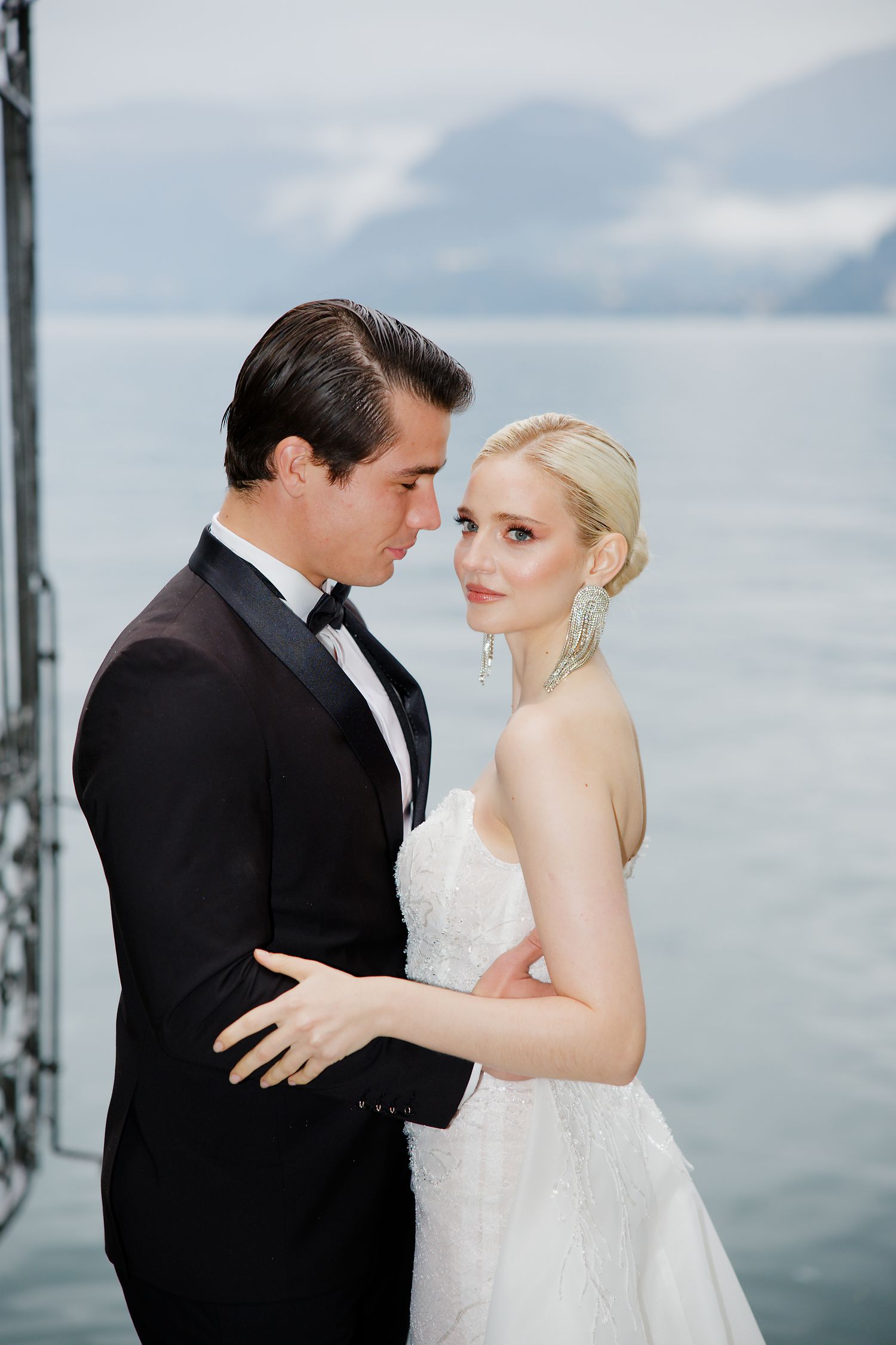 Elegant bride and groom embrace against a misty lake backdrop in formal wedding attire.