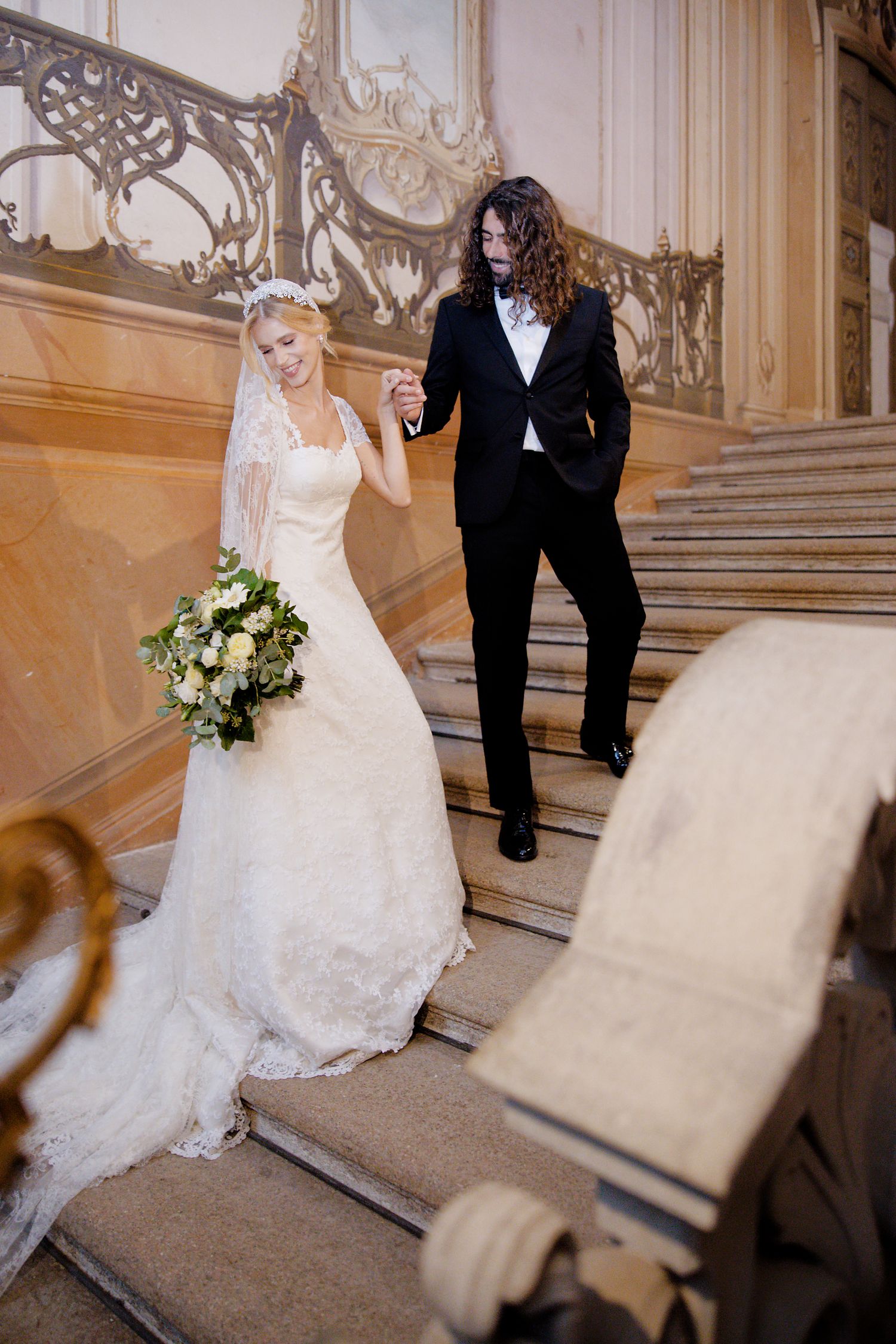 A bride in a white lace gown and groom descend an ornate marble staircase in an elegant historic venue.