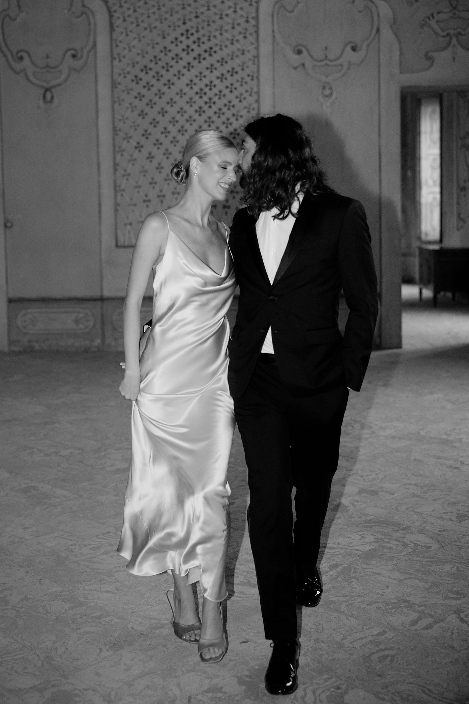 Black and white portrait of couple in formal attire walking together in ornate ballroom.