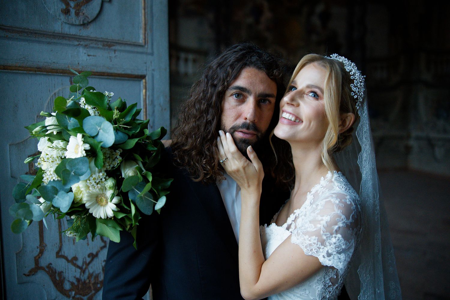 Romantic wedding portrait featuring couple with white bouquet against blue vintage door.