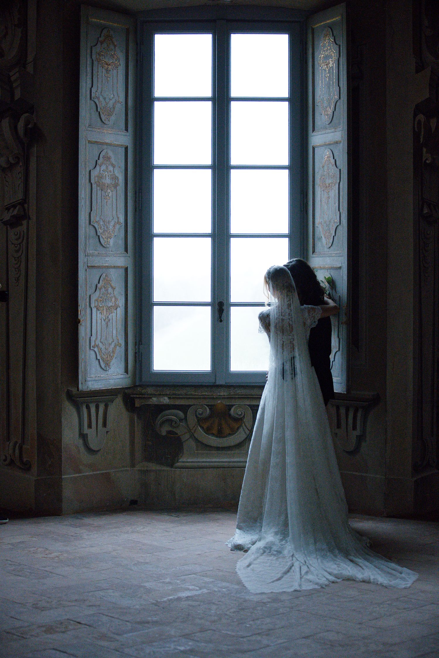 A figure in a flowing white wedding gown stands silhouetted against a tall ornate window in a historic interior.