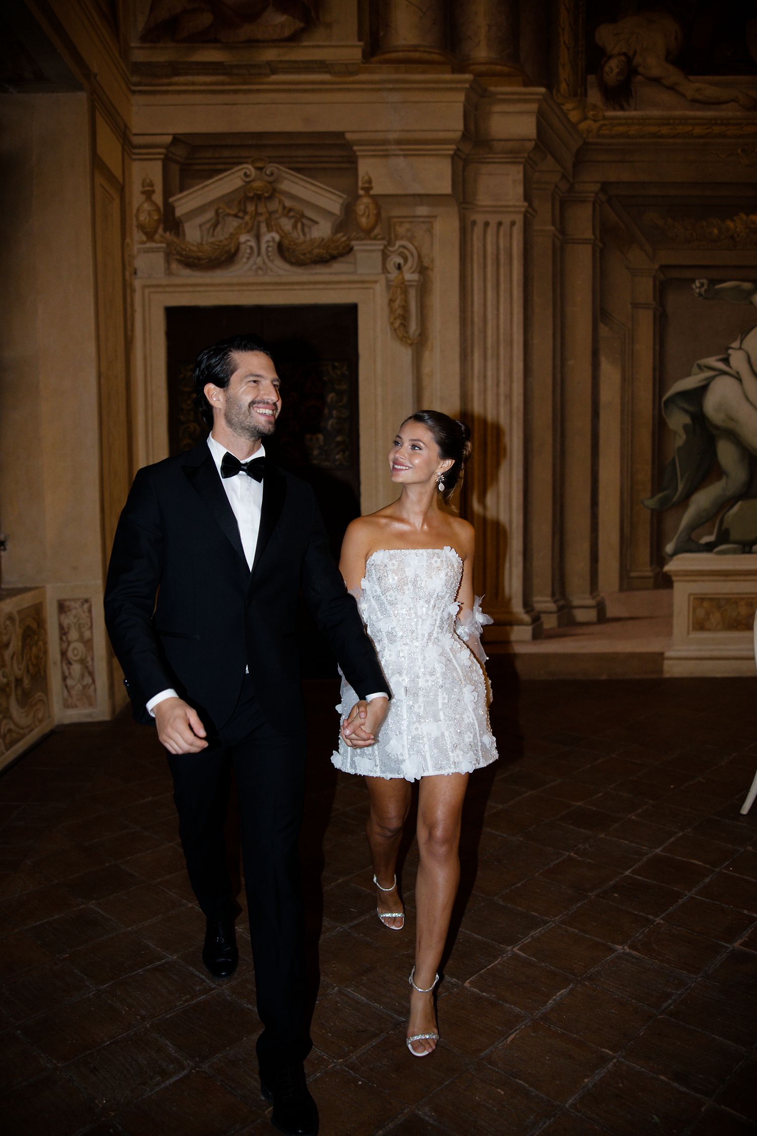 A couple in formal attire walks through an ornate hall with classical architecture and stone columns.