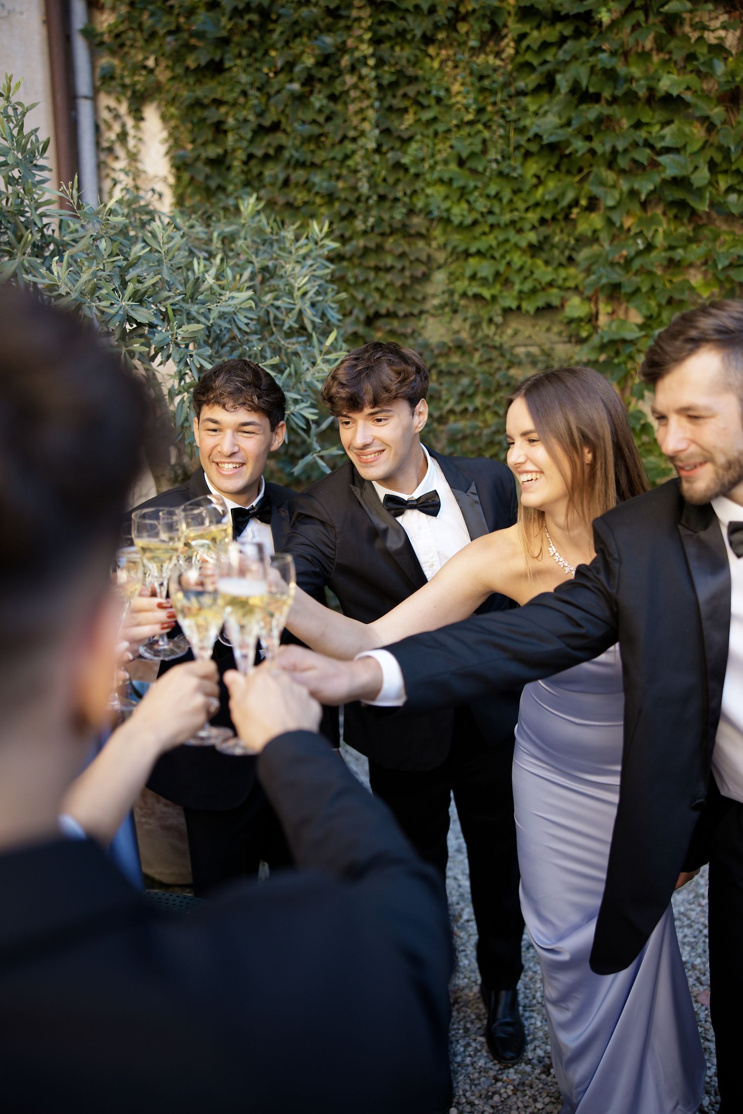 Wedding guests in formal attire toasting with champagne glasses in front of an ivy-covered wall.