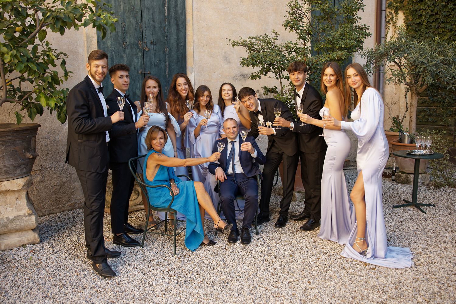 Wedding guests in formal attire gather for a group photo in an outdoor courtyard.