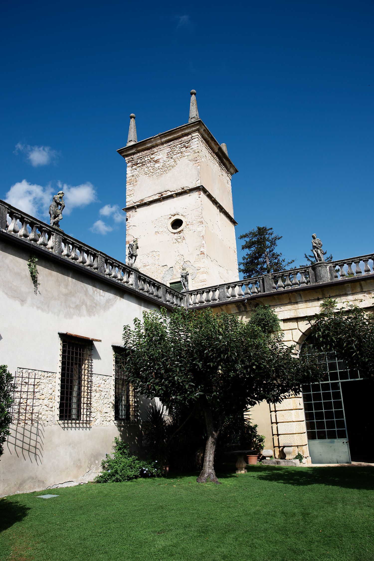 White stone tower with classical architectural details rises against bright blue sky in a manicured garden.