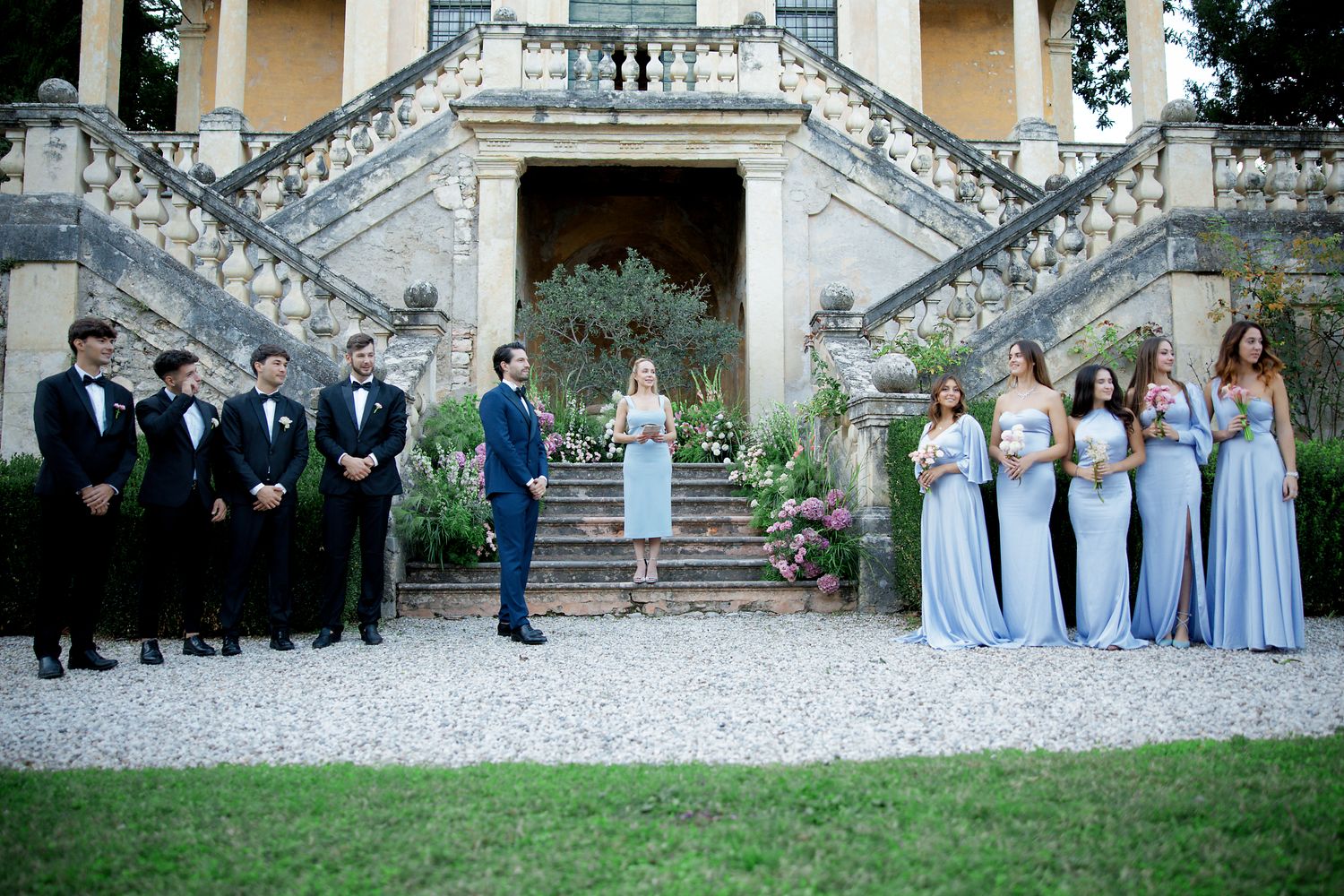 Wedding party poses on grand staircase with bridesmaids in blue gowns and groomsmen in black tuxedos.