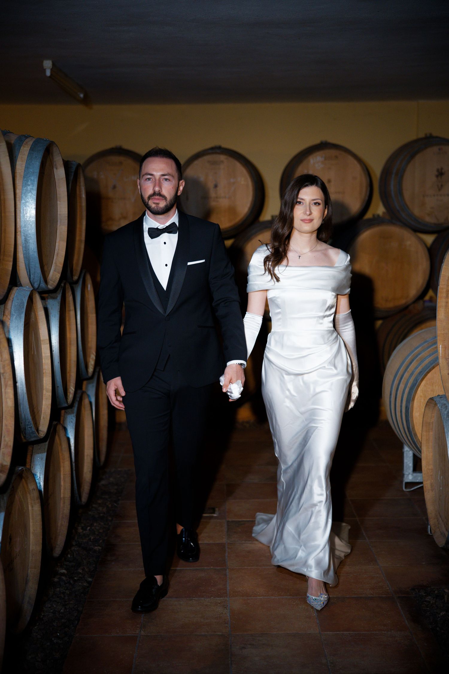 A newlywed couple walks through a rustic wine cellar lined with wooden barrels.