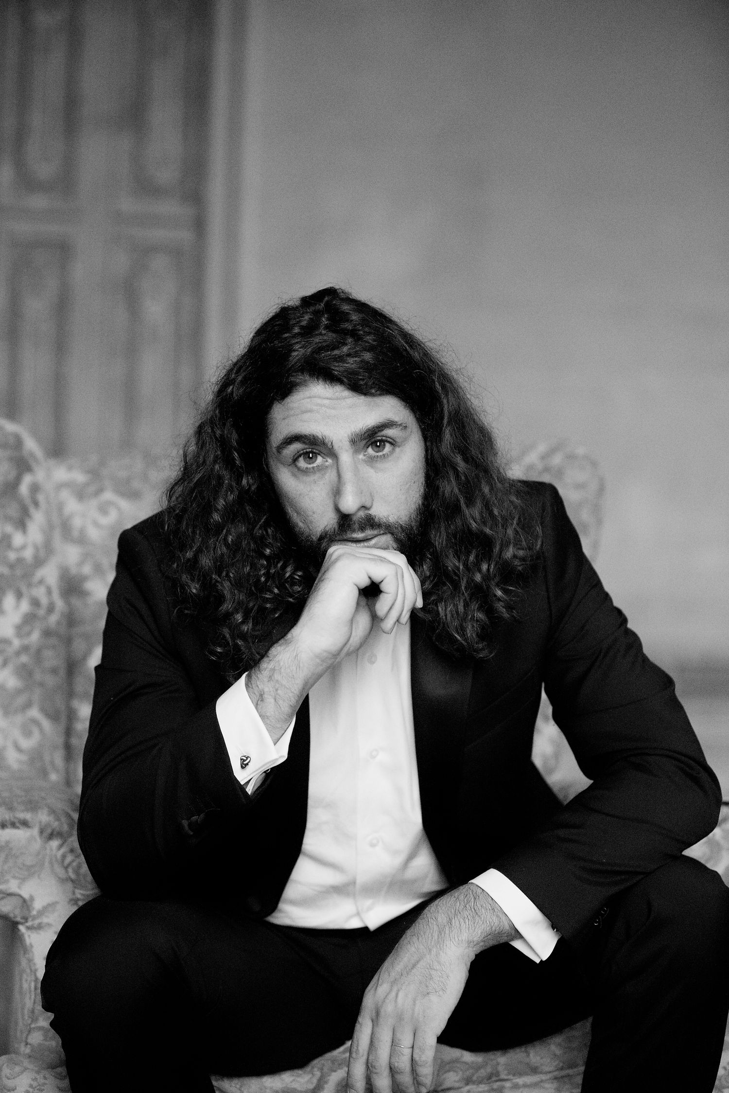 Portrait of man in formal suit with long dark hair seated in elegant setting.