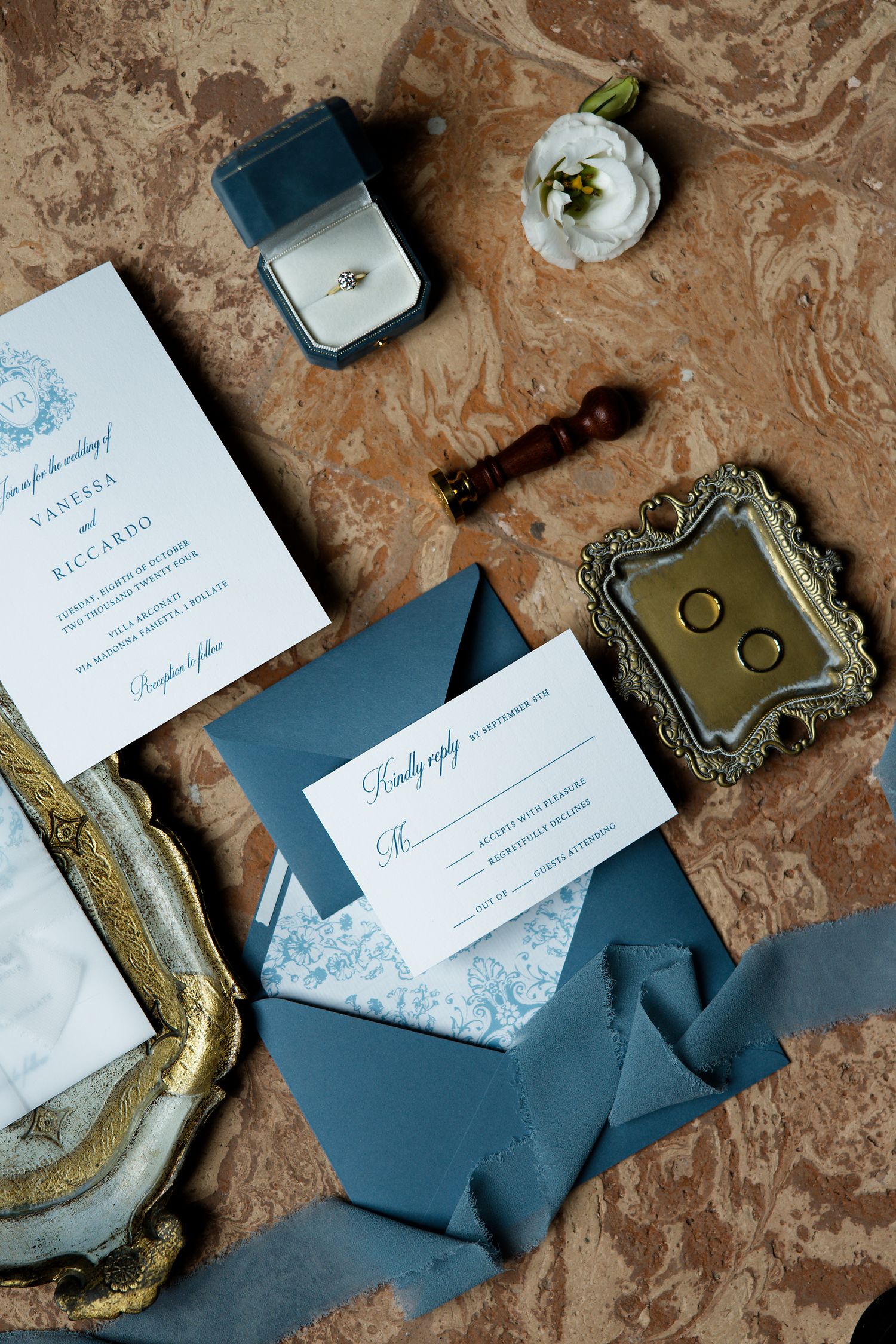 Wedding stationery set with blue envelopes, invitations, rings in box, and white flower on marble surface.