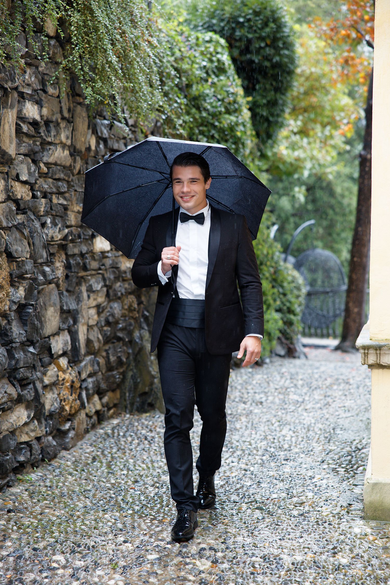 A person in a black tuxedo holding an umbrella walks along a stone wall path in an elegant garden setting.