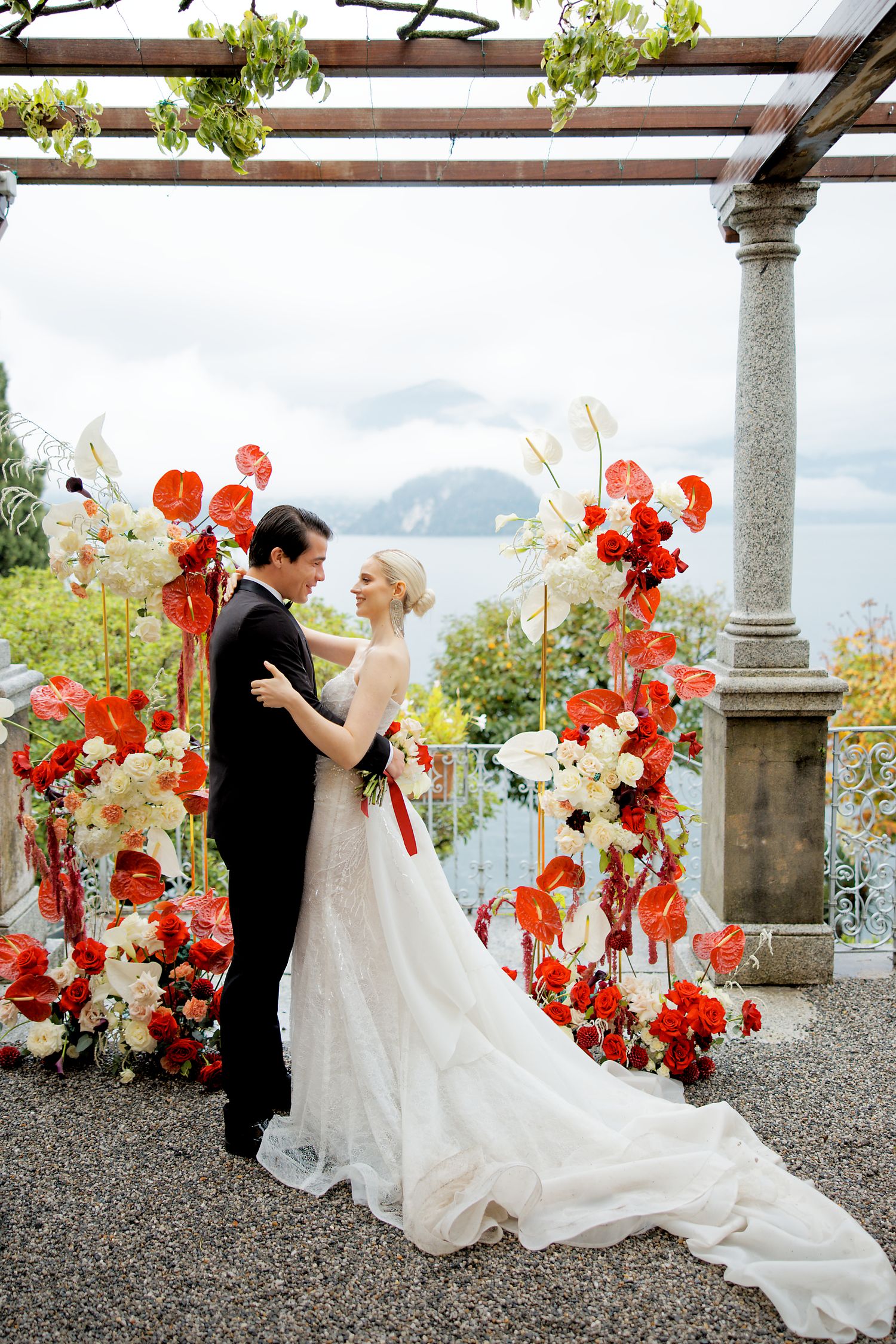 A romantic wedding portrait under a pergola decorated with vibrant red floral arrangements.