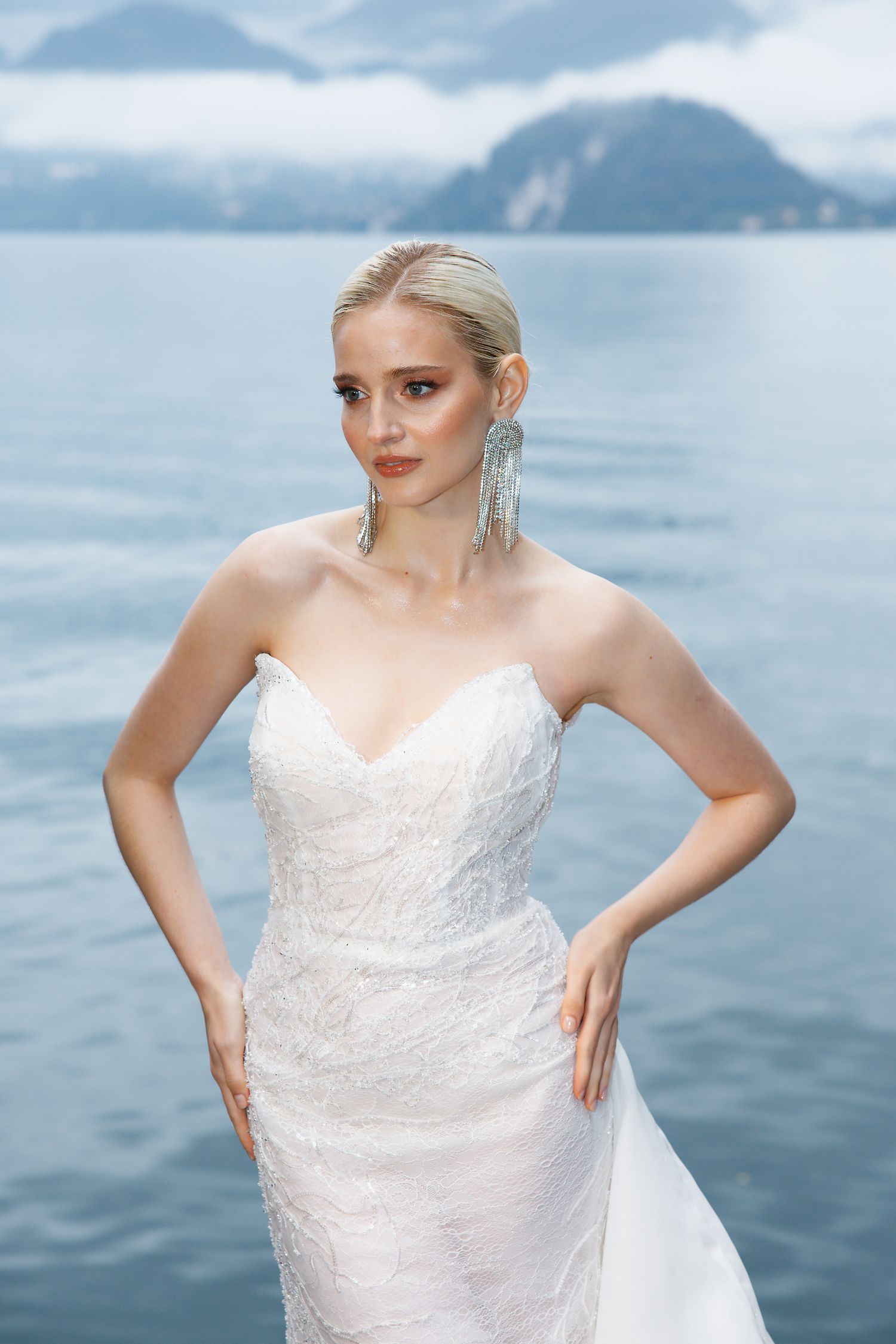 Bride in white lace gown poses against backdrop of mountain lake.