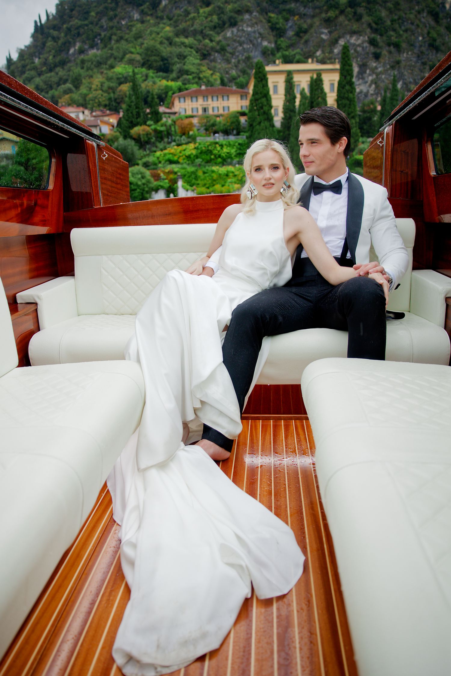 Newlyweds relax on luxurious white leather seats of wooden boat with Italian villa in background.
