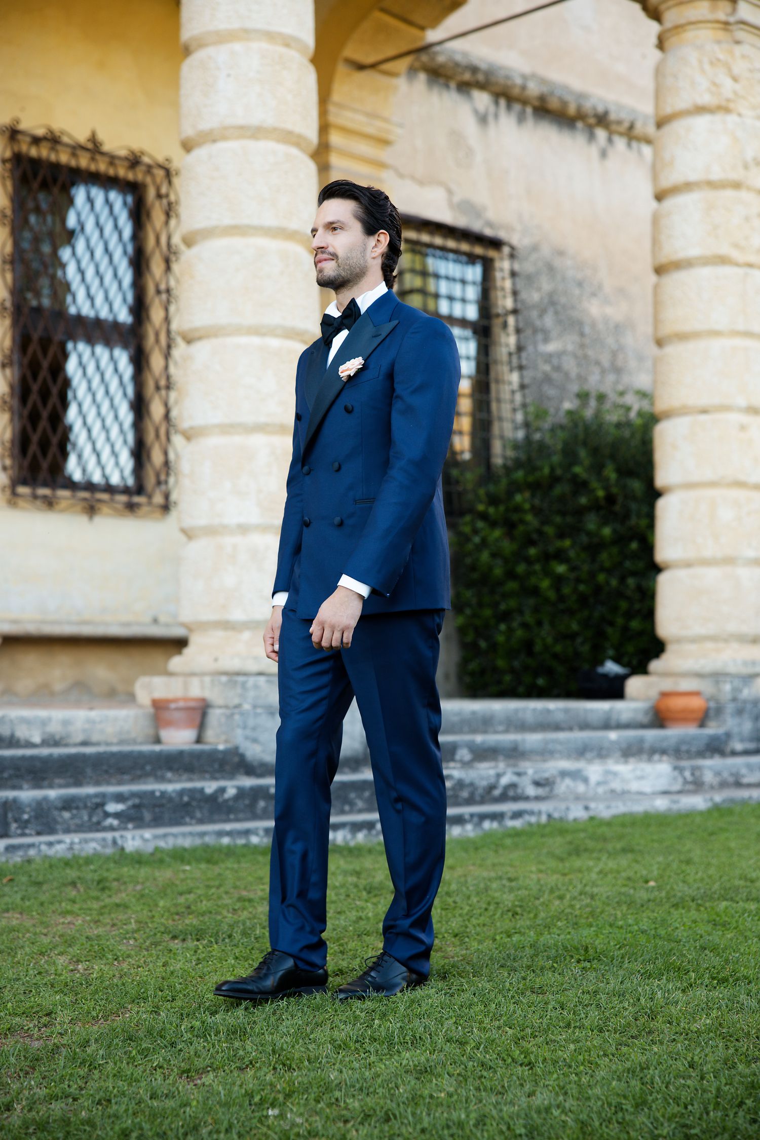 A groom in a tailored navy blue suit stands on manicured grass outside a historic stone building with columns.