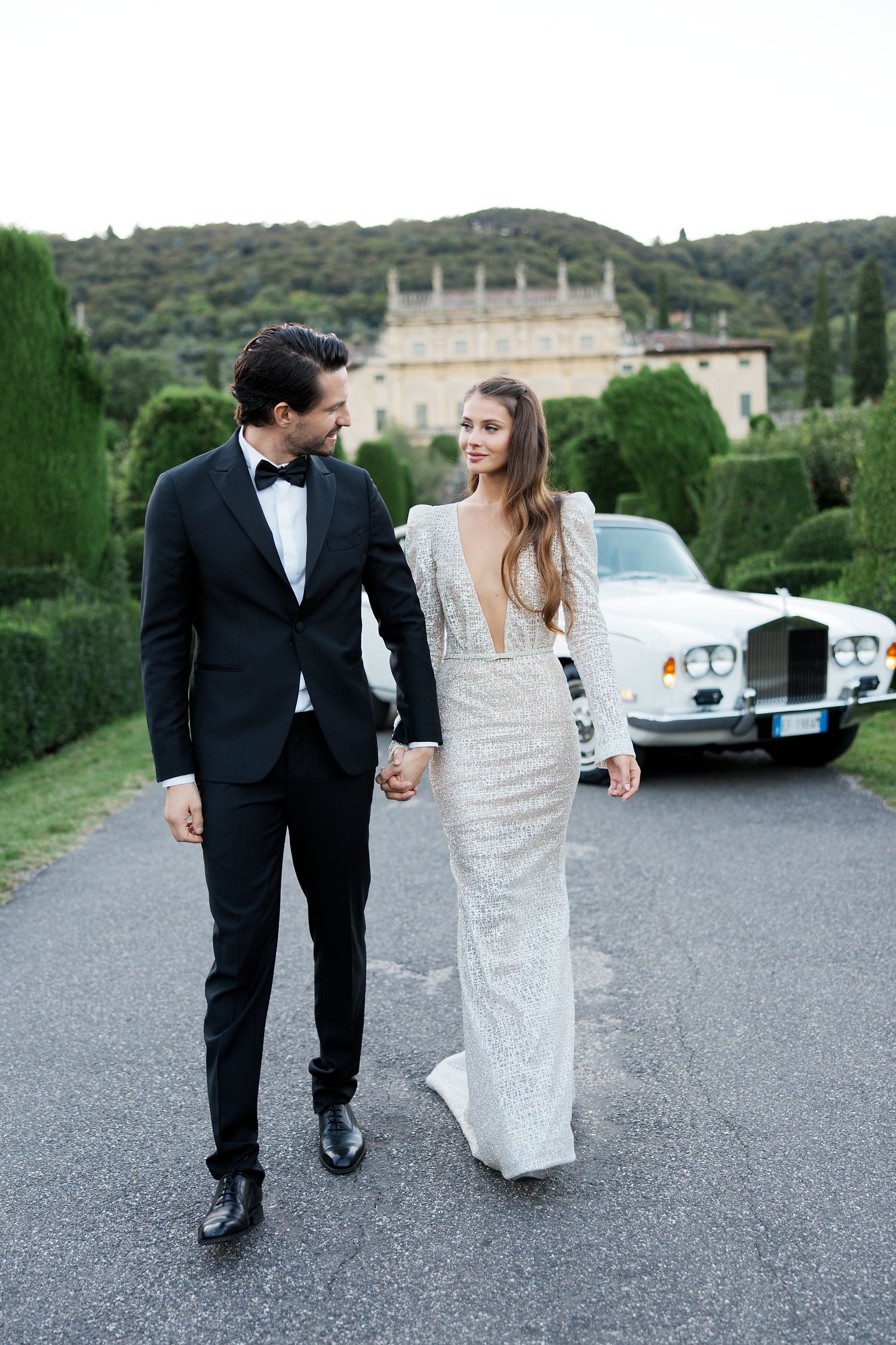 Newlyweds walking hand in hand near a classic car with grand villa in background.