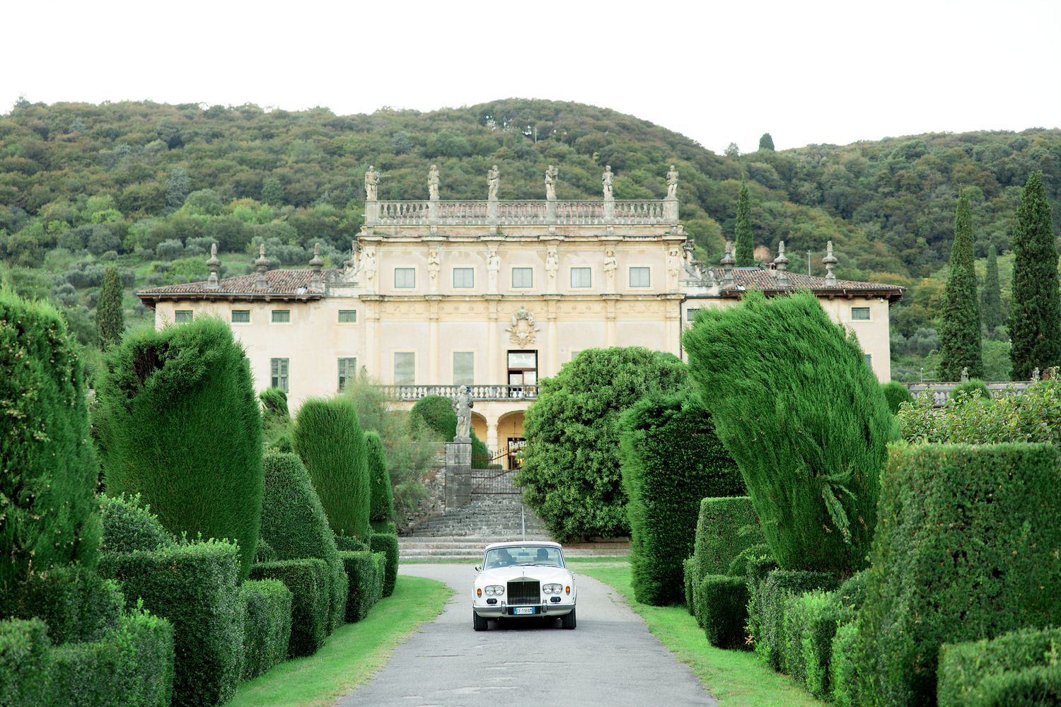 A vintage car drives down a formal garden path towards an elegant Italian villa with manicured topiary hedges.