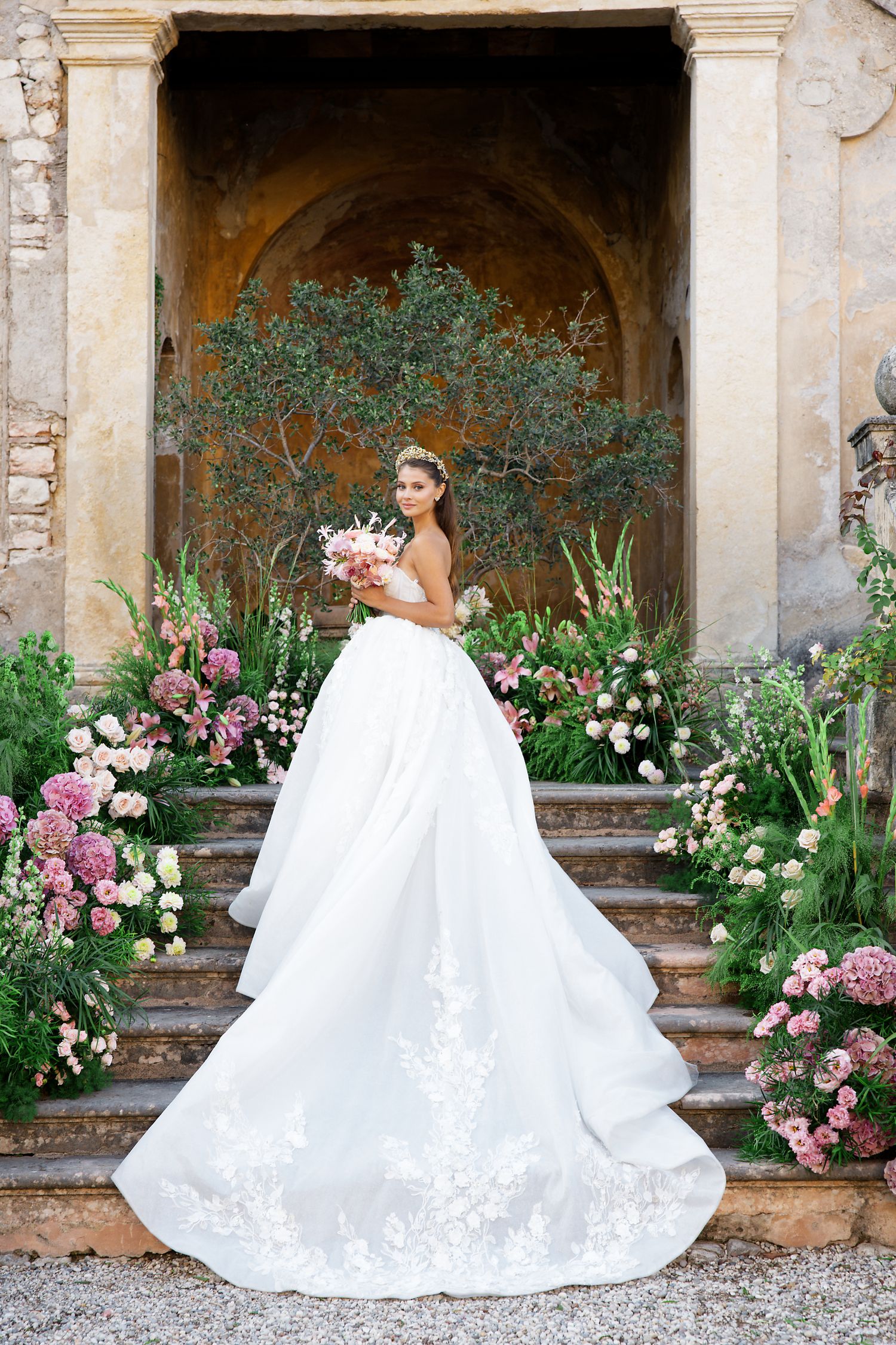 Bride in ornate white gown photographed under stone archway with pink and white floral garden arrangements.