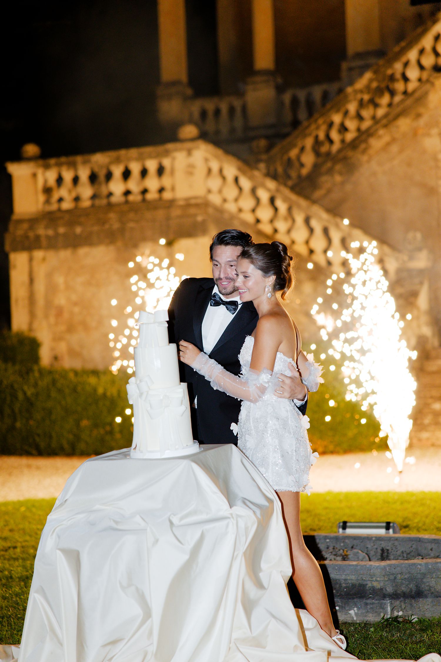 Wedding couple poses with sparkler fountains and white cake at night.