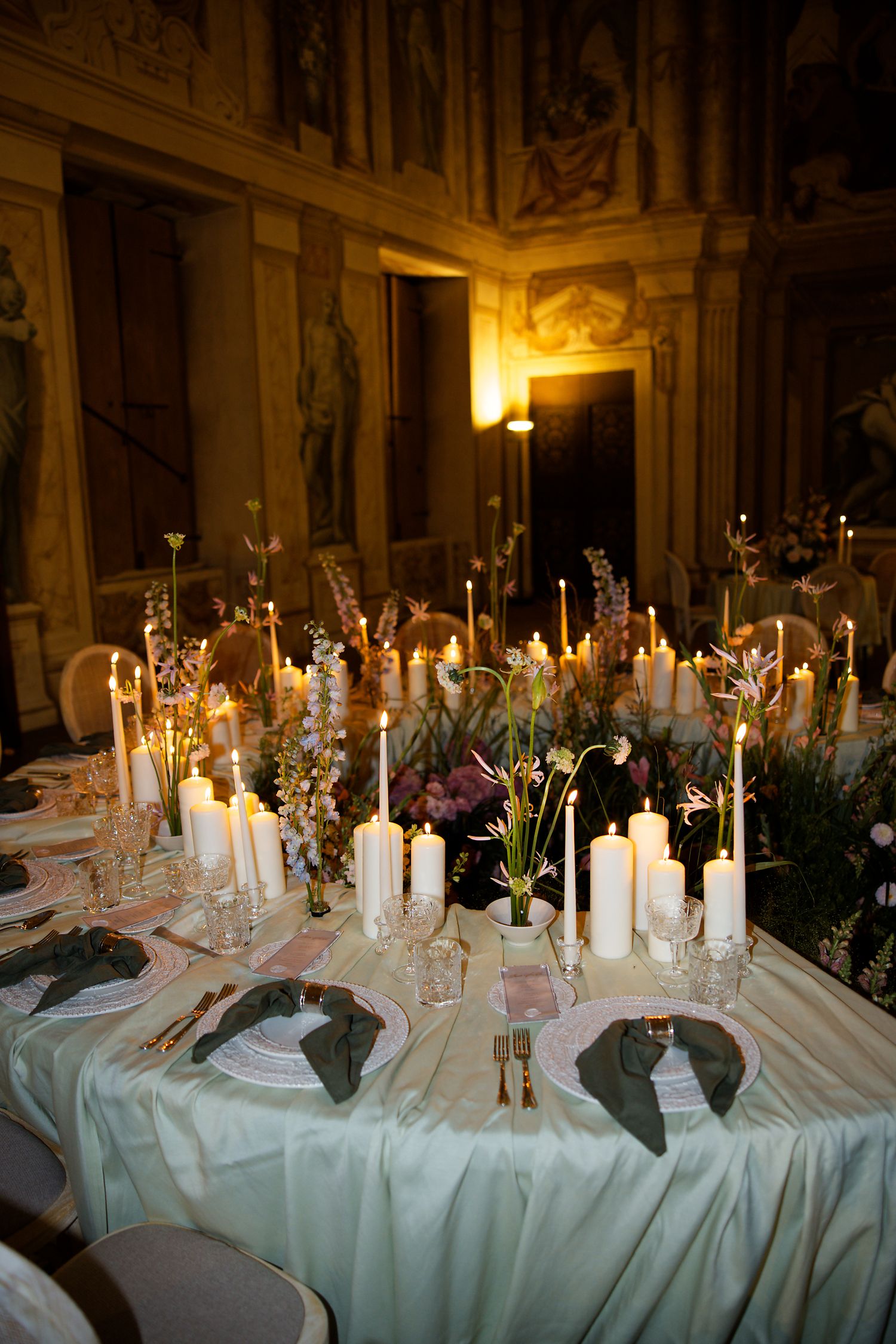 Romantic candlelit wedding table setting with white tapers and floral centerpieces in an ornate historic hall.