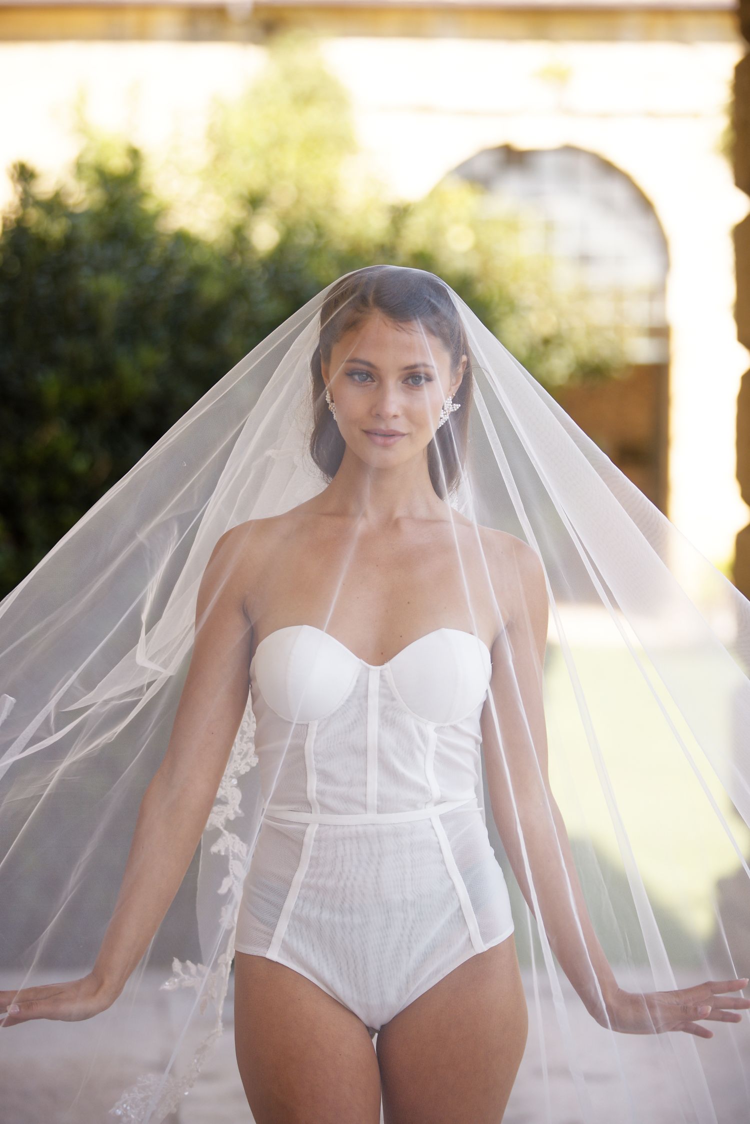 Bride posing in a white bridal bodysuit and flowing cathedral veil in natural sunlight.
