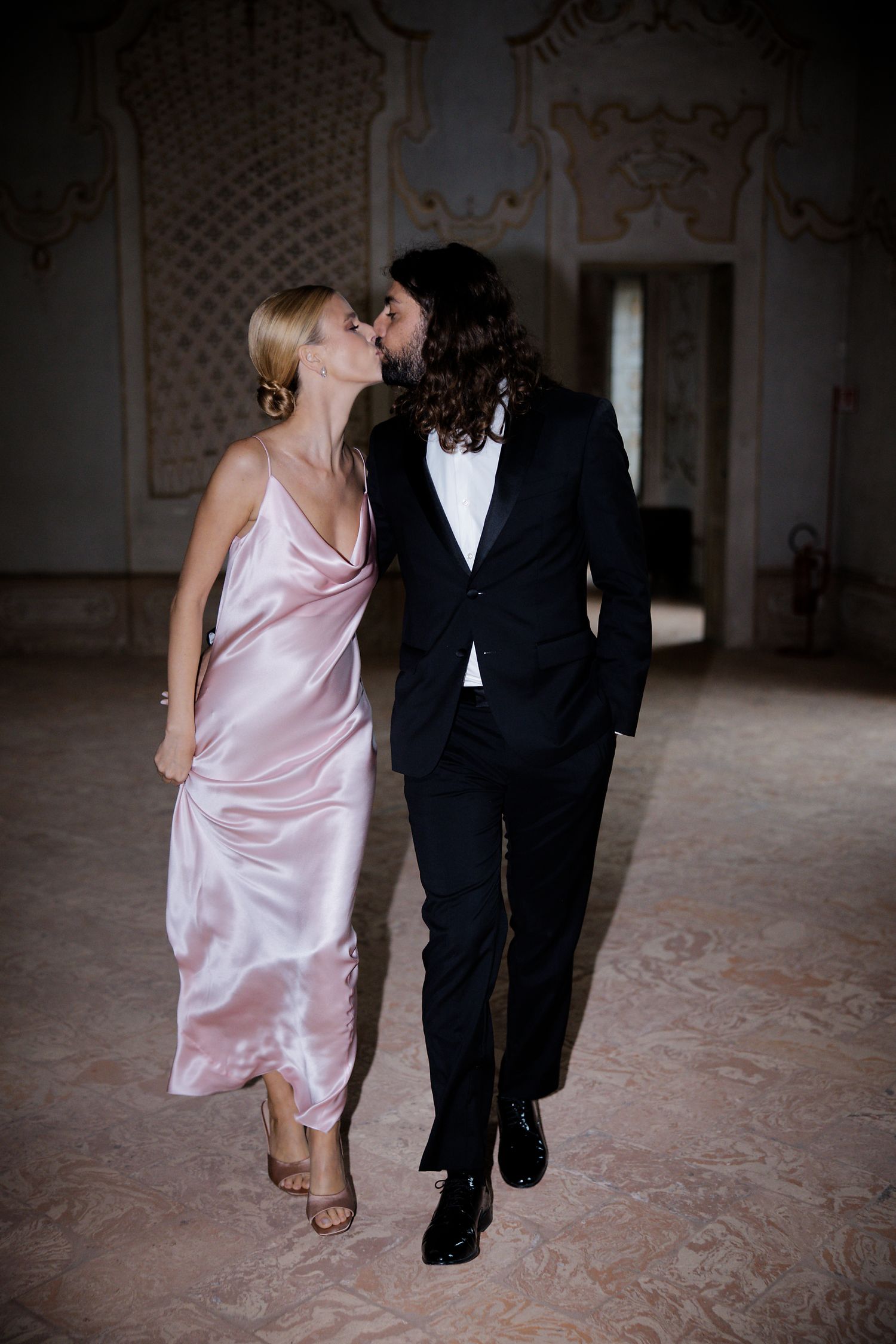 A couple in evening wear share an intimate moment in an ornate room with decorative wall patterns.