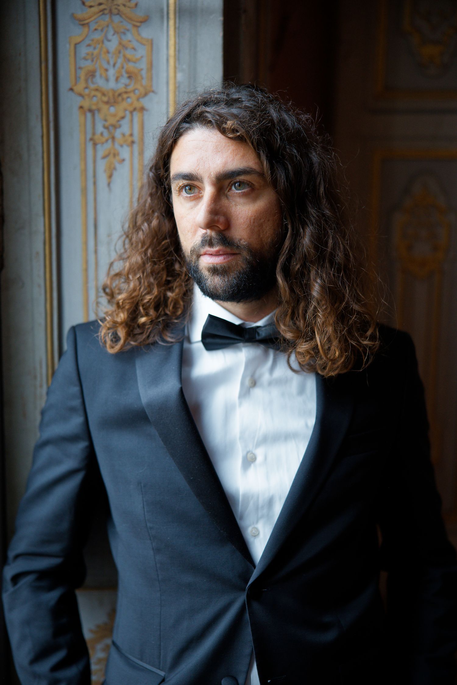 Person in black tuxedo and bowtie with long wavy hair poses against decorative wall with gilded details.