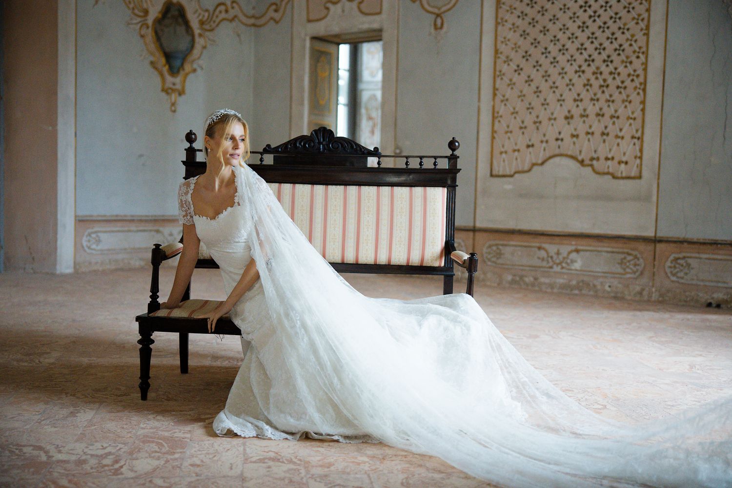 Bride in flowing white gown posed elegantly on antique chair in ornate room.