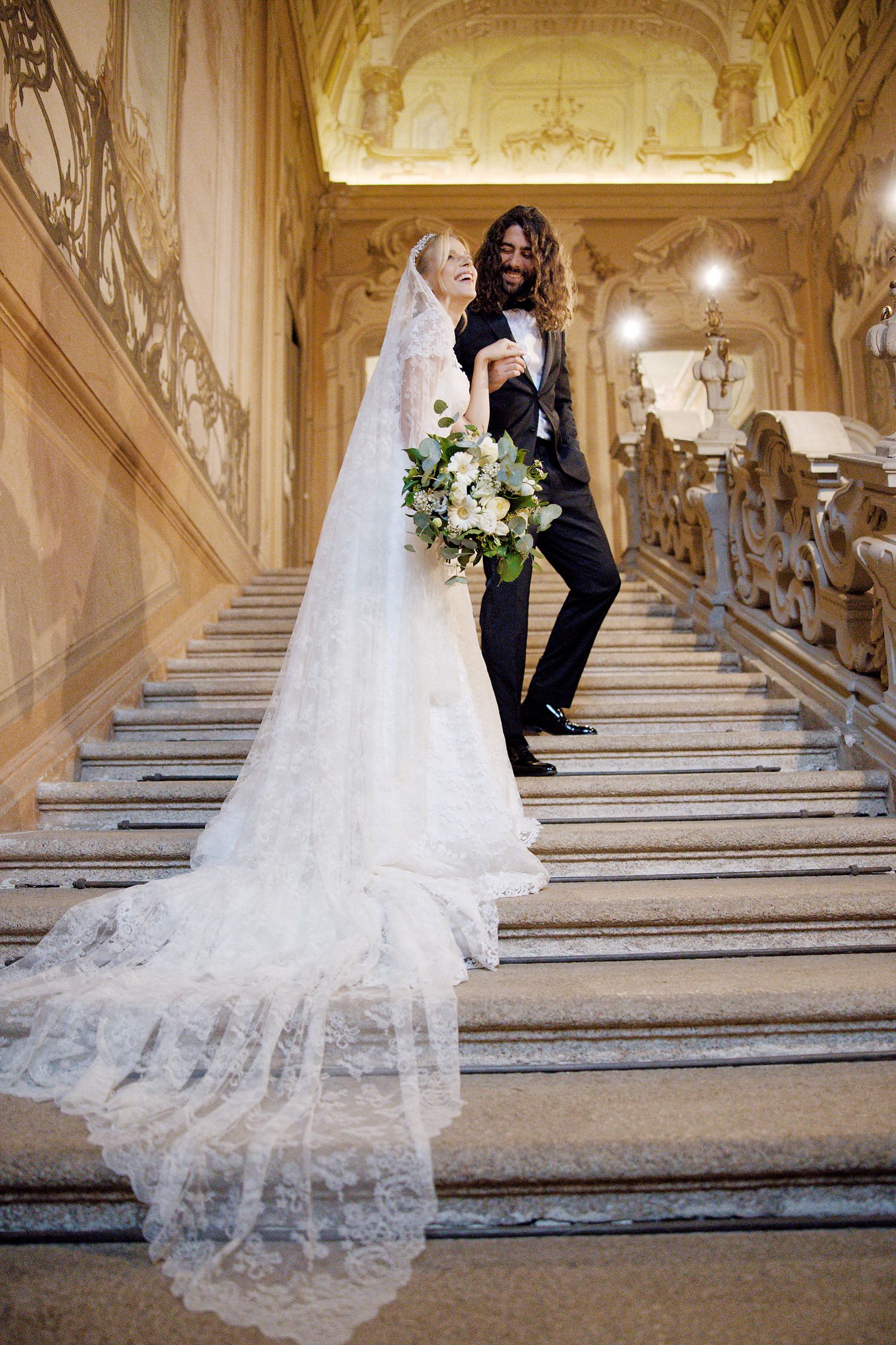 Wedding couple poses on ornate marble staircase with bride wearing flowing lace veil and holding white bouquet.