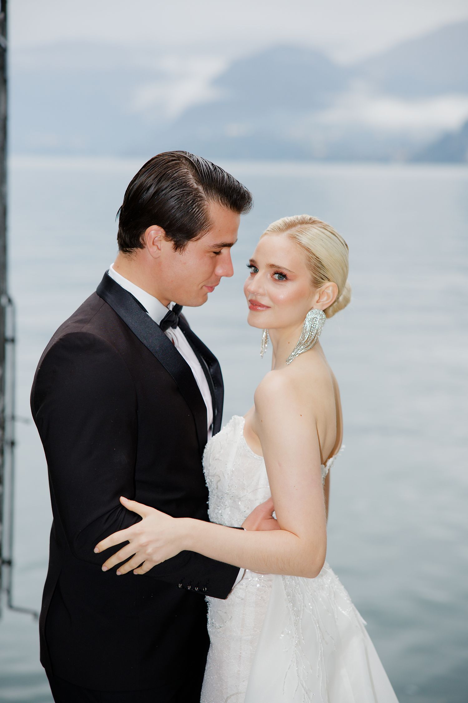 Elegant wedding couple embraces against a misty lake backdrop in formal attire.