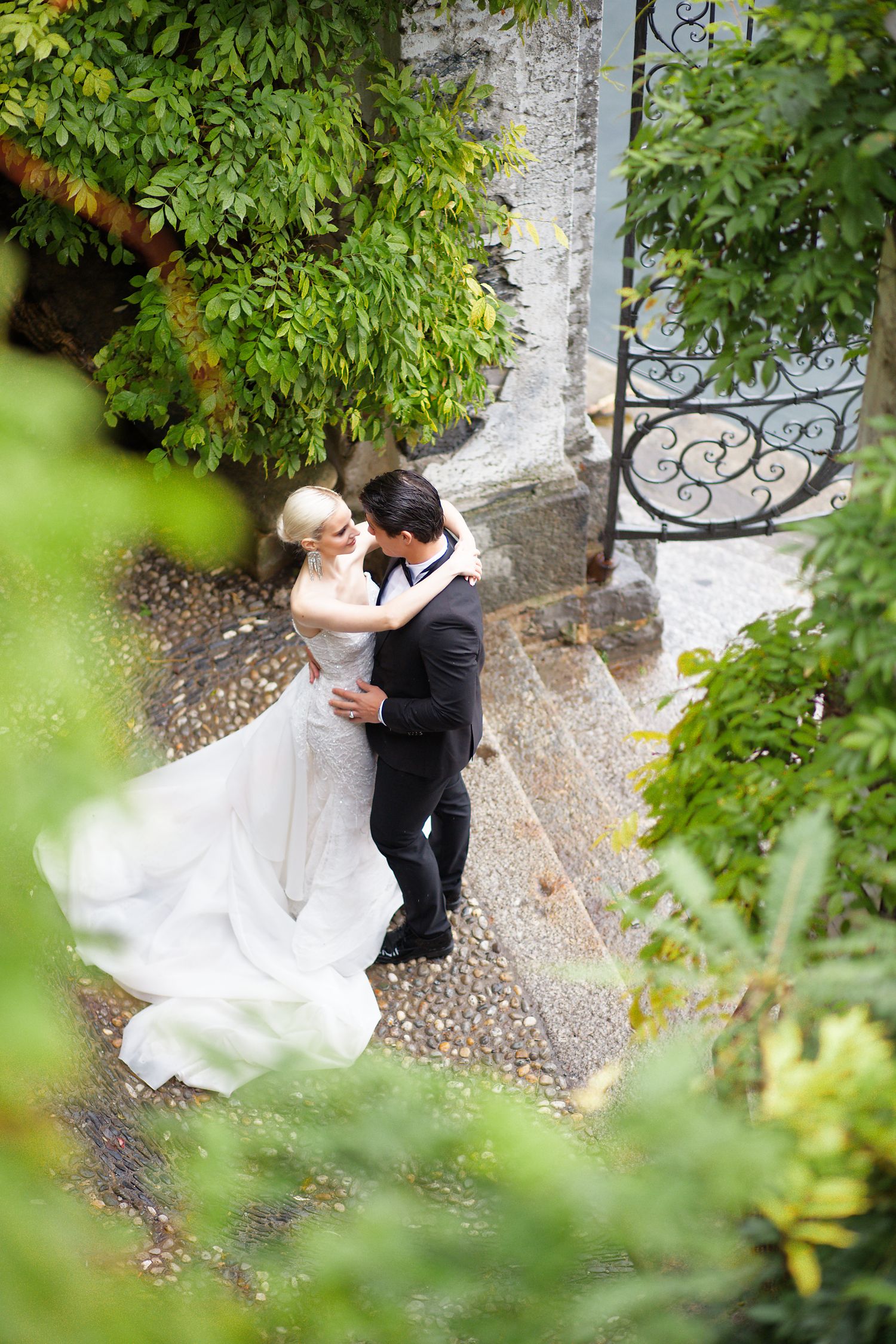 A romantic wedding couple embraces on a cobblestone path framed by lush green foliage and ornate iron gates.