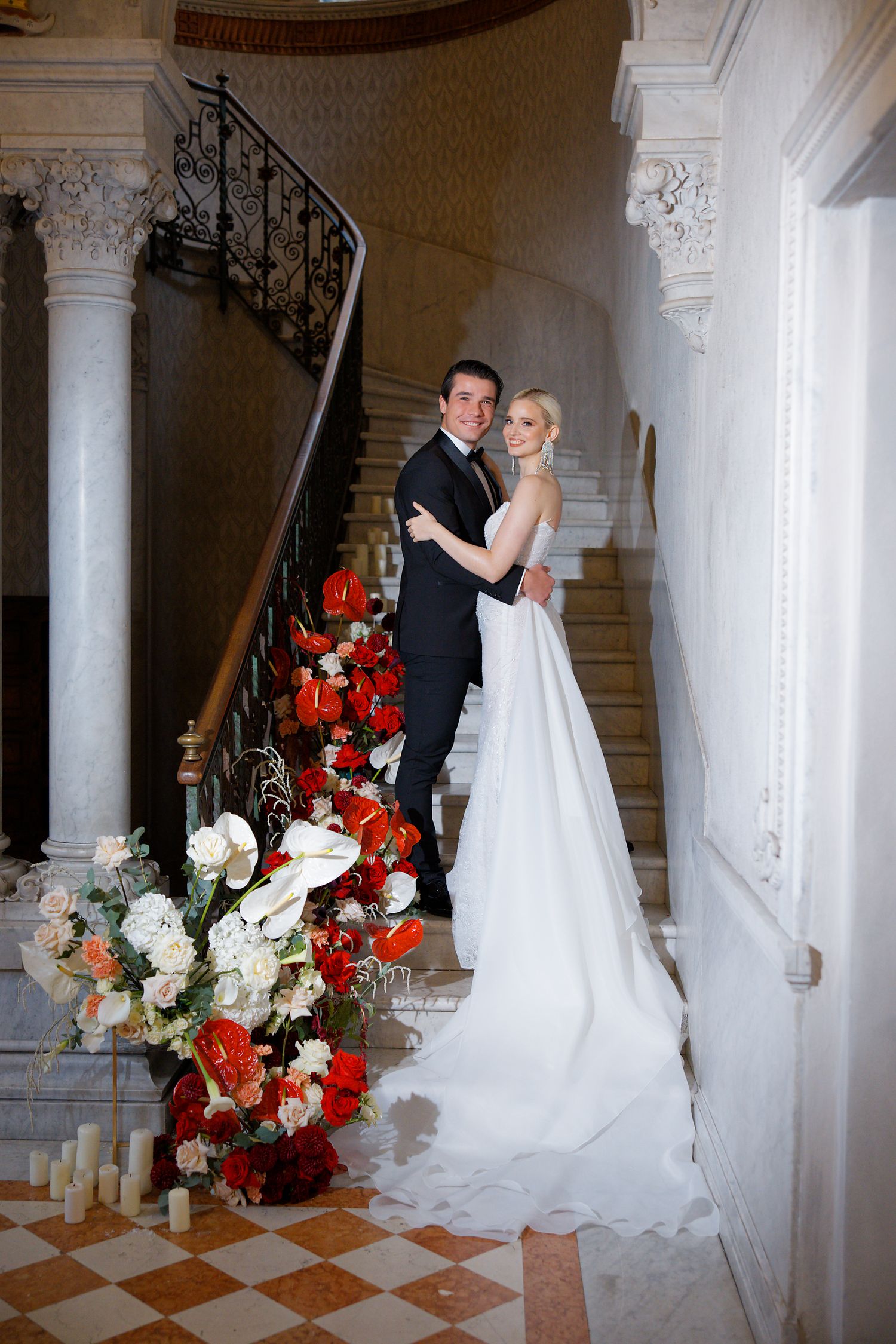 Elegant wedding couple poses on grand staircase decorated with red and white floral arrangements in historic mansion.