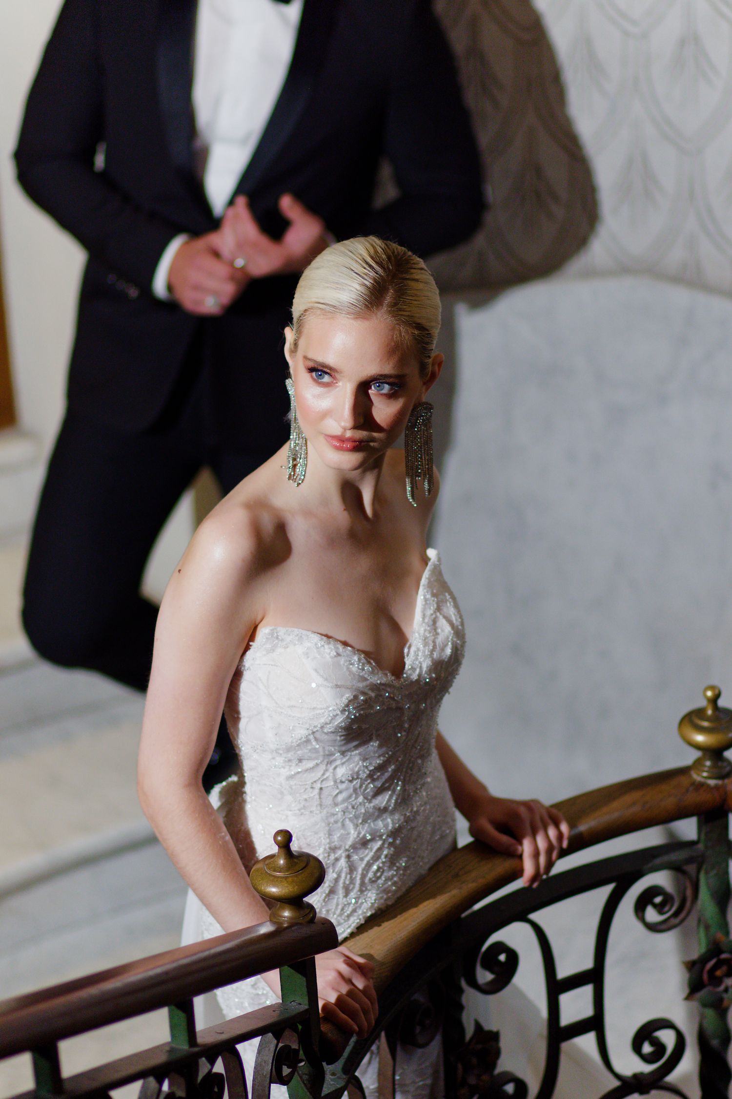 Elegant bride in white lace strapless gown poses on ornate staircase with dangling earrings and sleek updo.