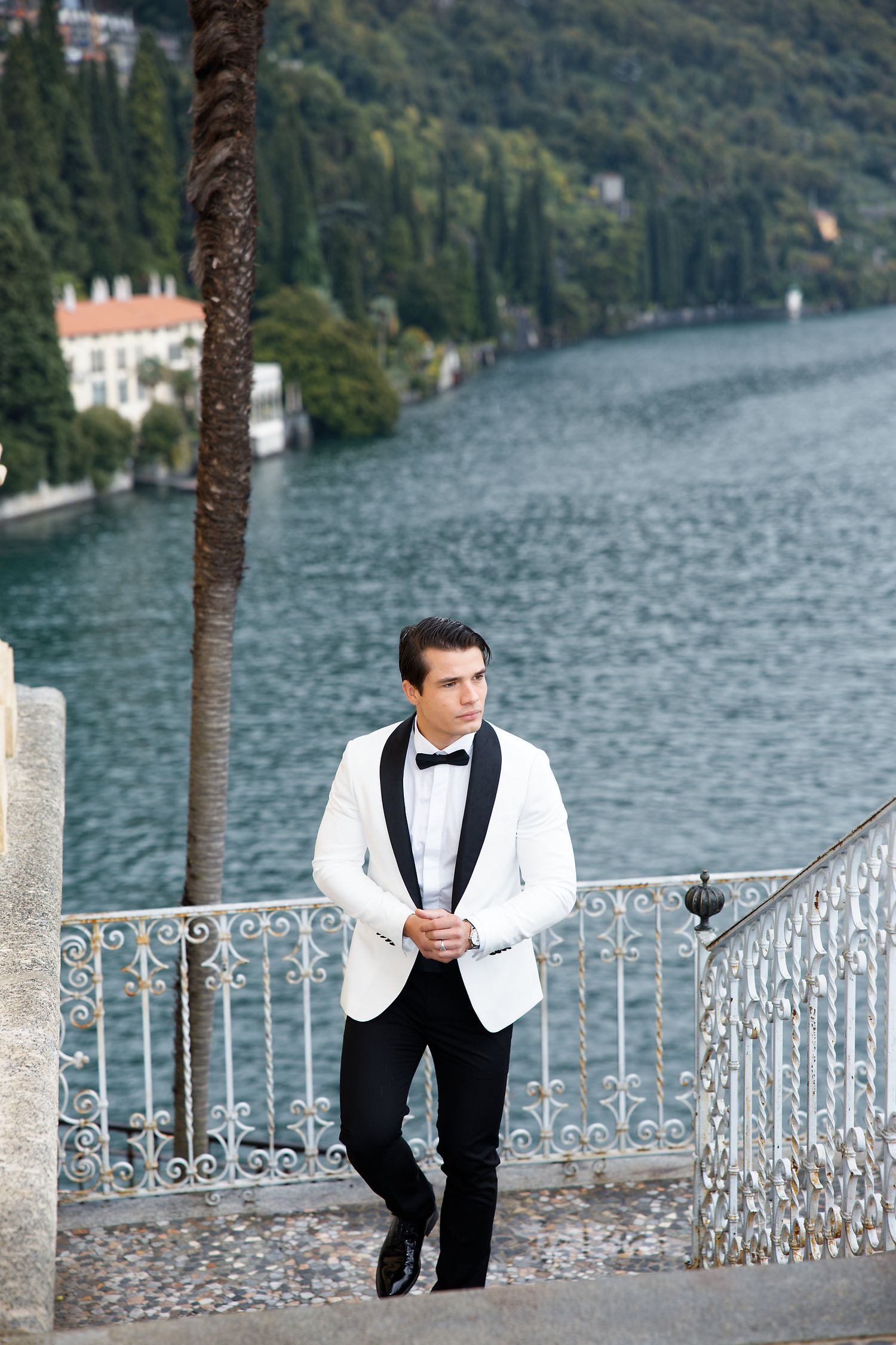 A person in a white dinner jacket and bow tie stands by an ornate railing overlooking a lakeside in Italy.