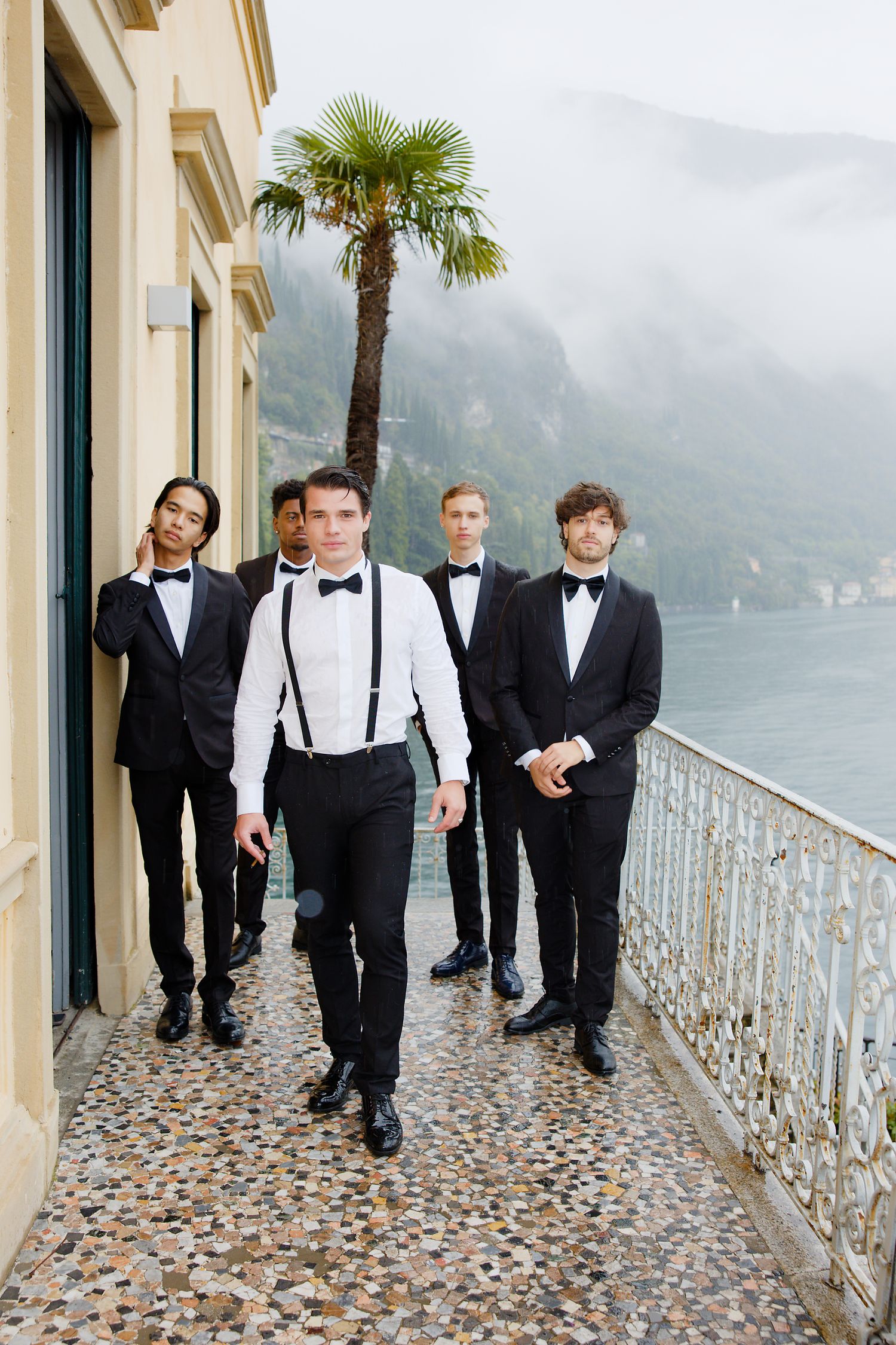 Group of groomsmen posing on a lakeside terrace with mountains and palm trees in the background.