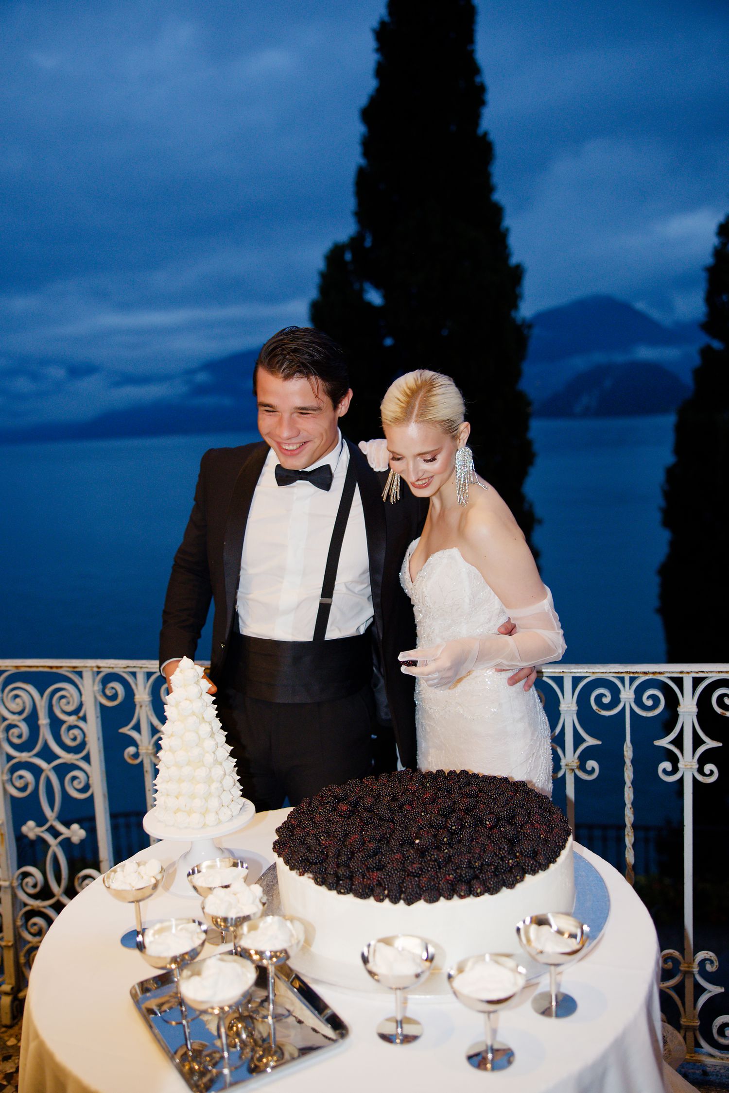 Elegant wedding couple poses with their two-tiered cake display on a lakeside balcony at dusk.