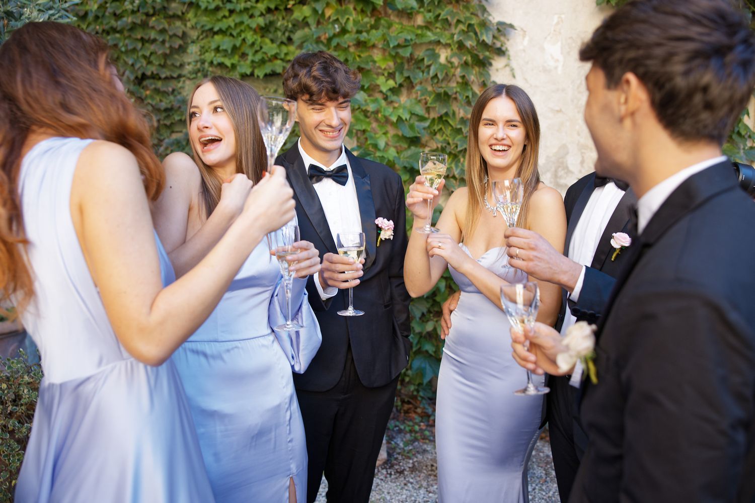 Guests in formal attire laugh and socialize while holding champagne glasses at an outdoor event against an ivy-covered wall.
