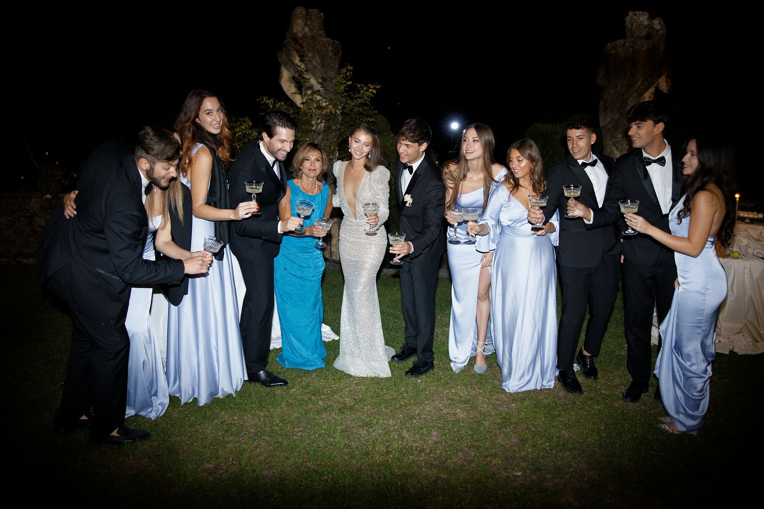 Wedding party guests in formal attire gather for an evening celebration on a dark lawn.