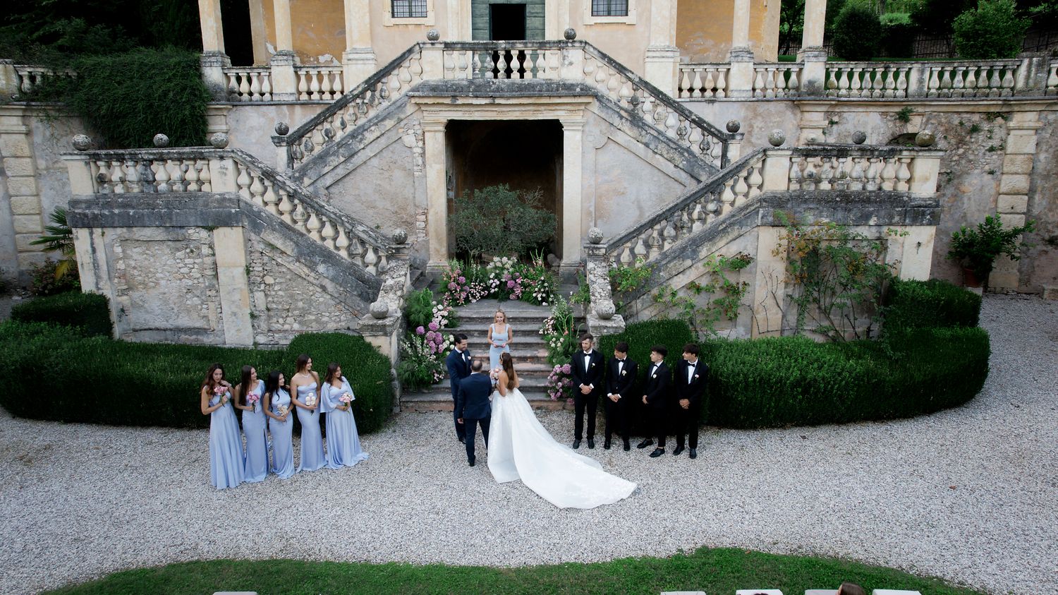 Wedding party poses symmetrically on grand stone staircase of Italian villa.