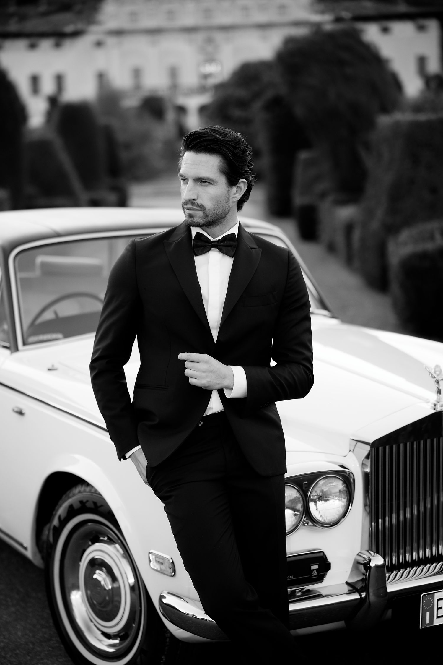 Classic black and white photo of groom in tuxedo leaning against vintage Rolls Royce car.