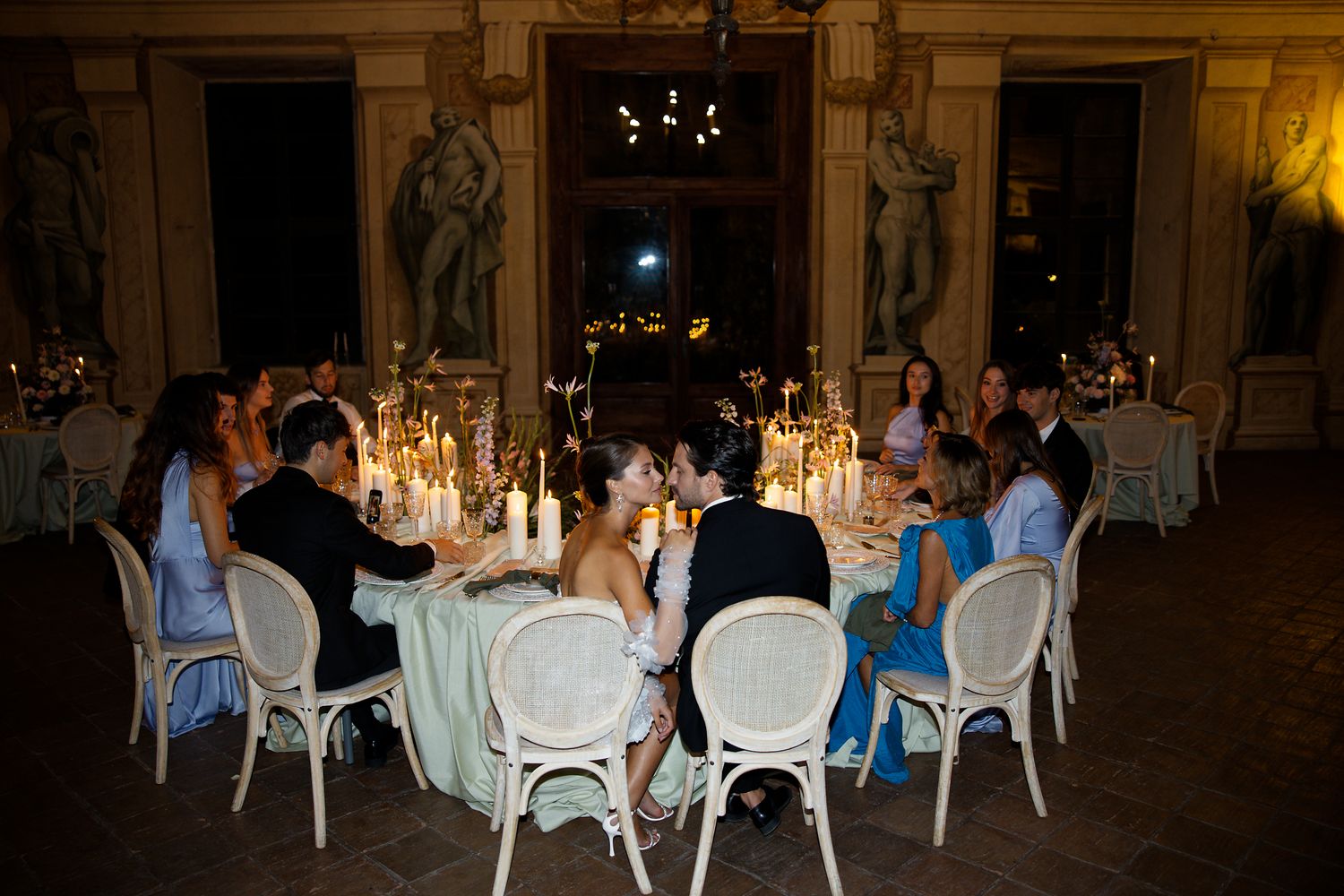 An elegant wedding reception dinner table setting with candles and white chairs in a grand historic hall.