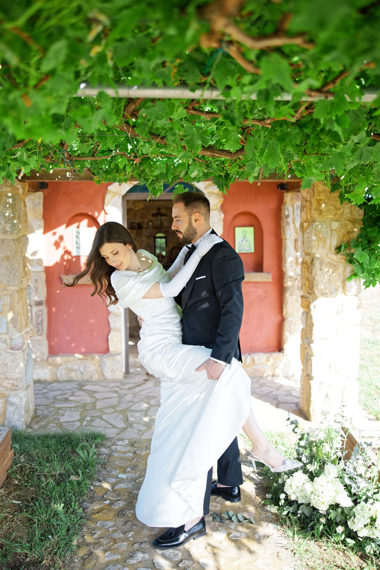 Romantic wedding couple embraces under green foliage near a rustic stone and coral colored wall.